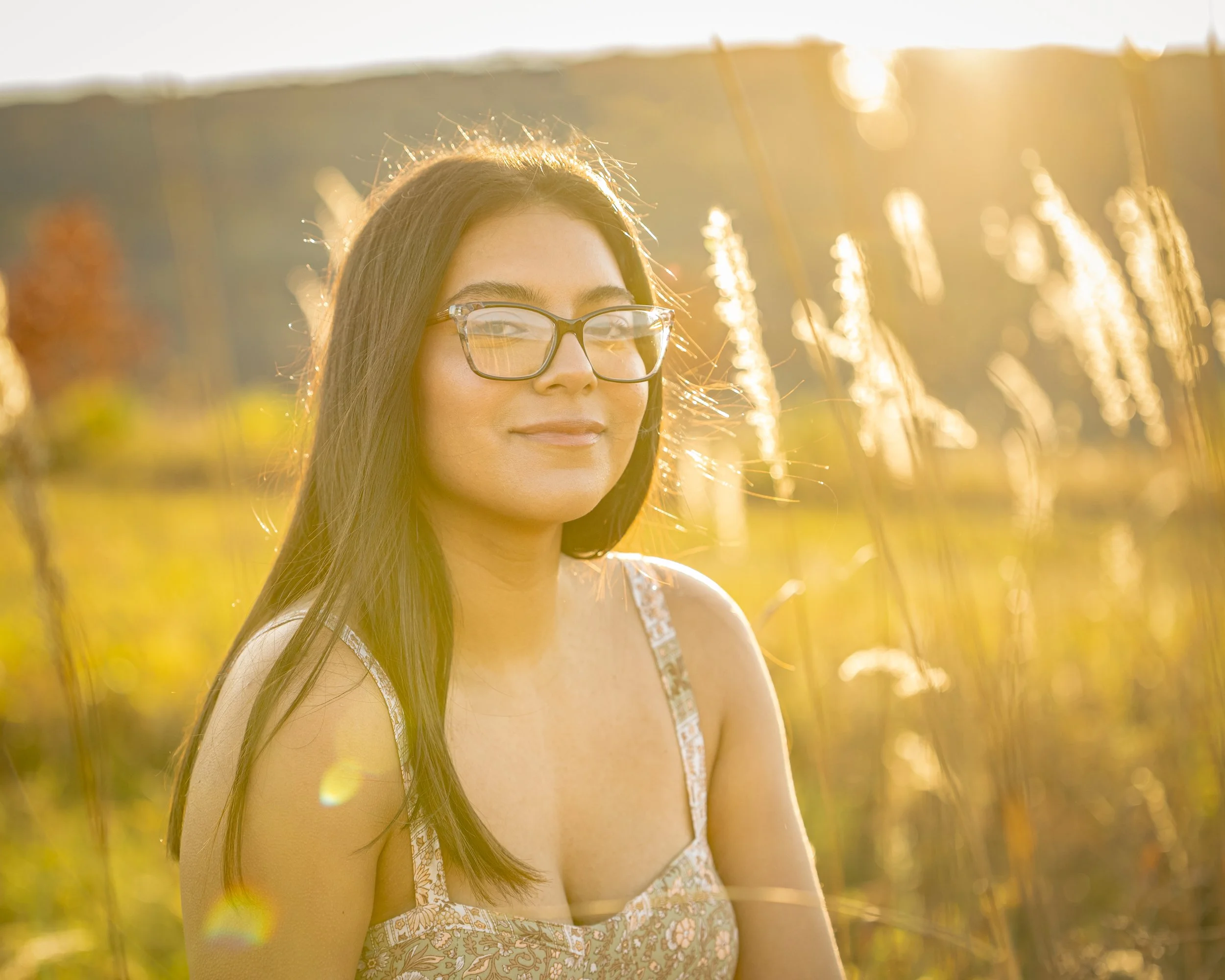 Young woman with long dark hair and glasses smiling in a sunny field with tall grass.