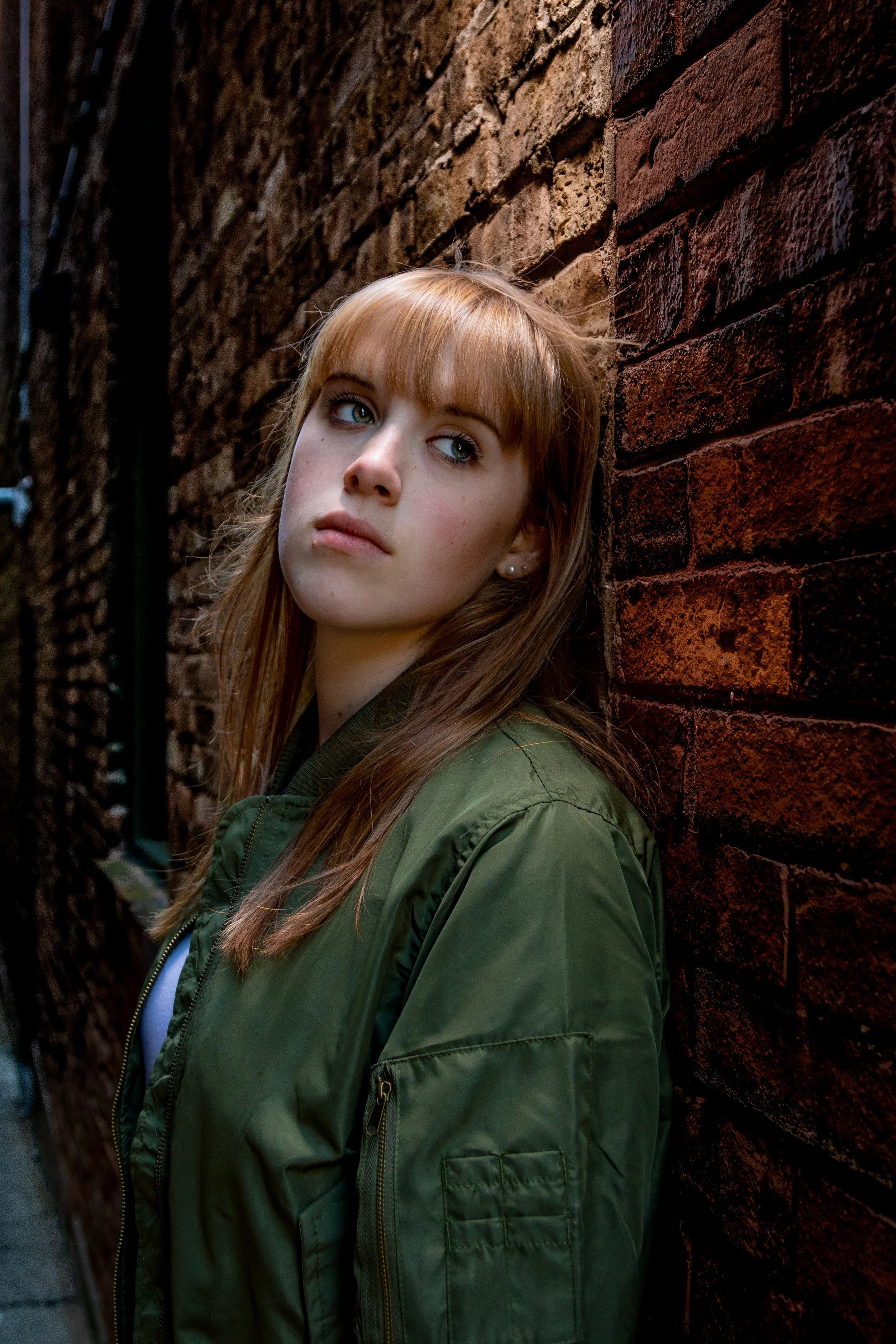 Person with long hair and green jacket leaning against a brick wall, looking thoughtful.
