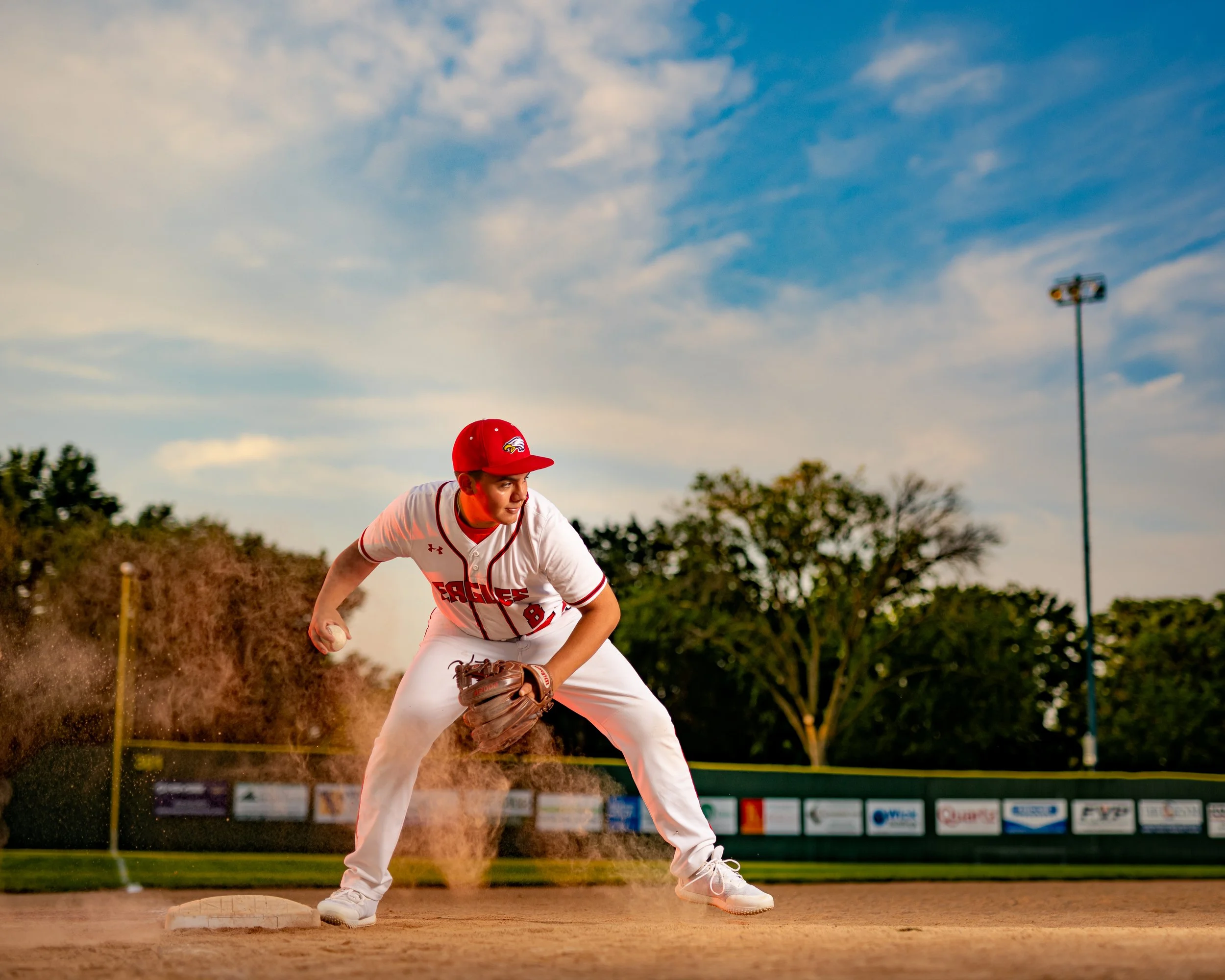 Baseball player fielding infield, wearing red and white uniform and cap, with glove and ball near a base, on a dirt field. Trees and advertisements in the background under a blue sky.