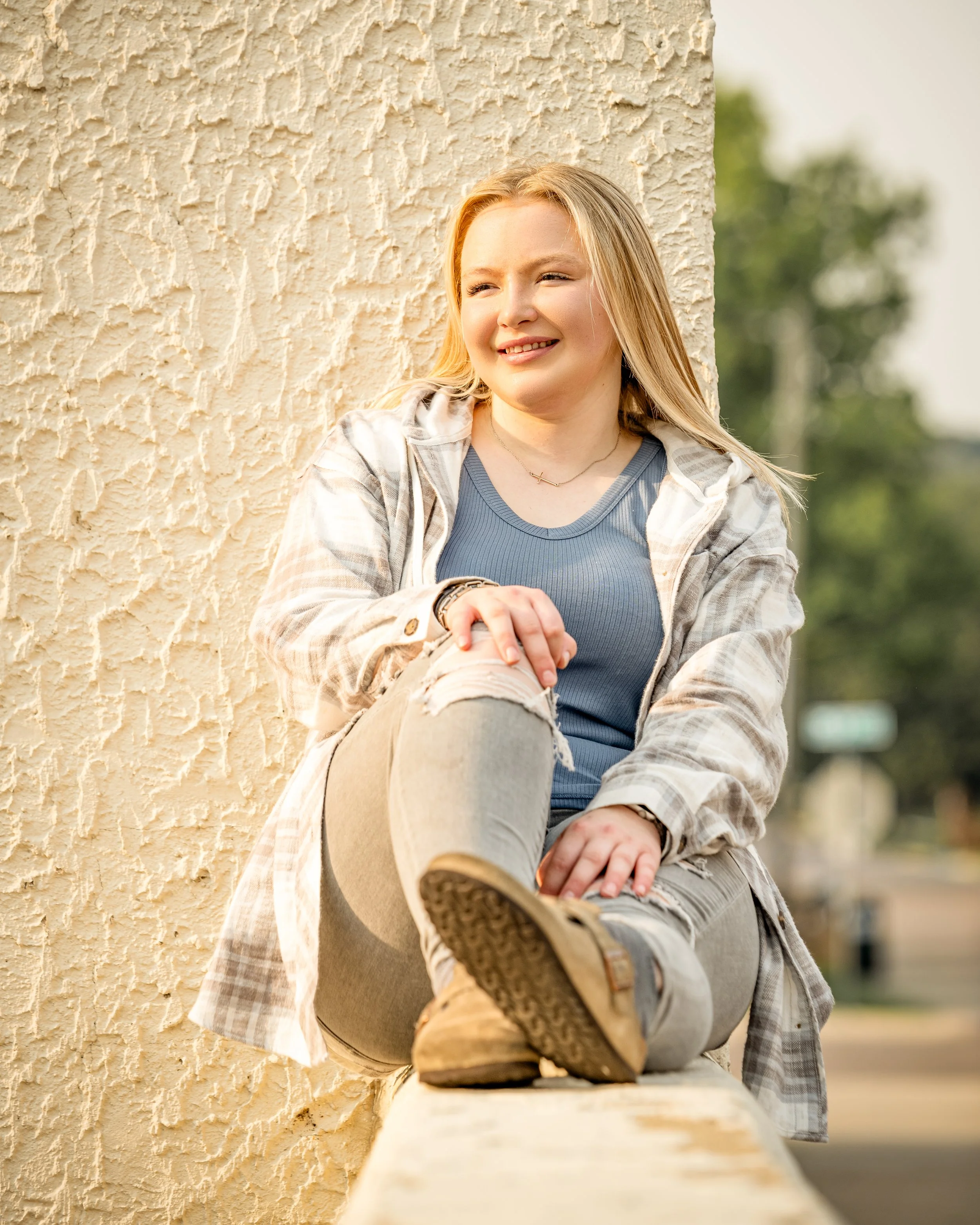 A person with long blonde hair sitting outdoors in casual attire, wearing ripped jeans, a blue top, and a plaid shirt, leaning against a textured wall.