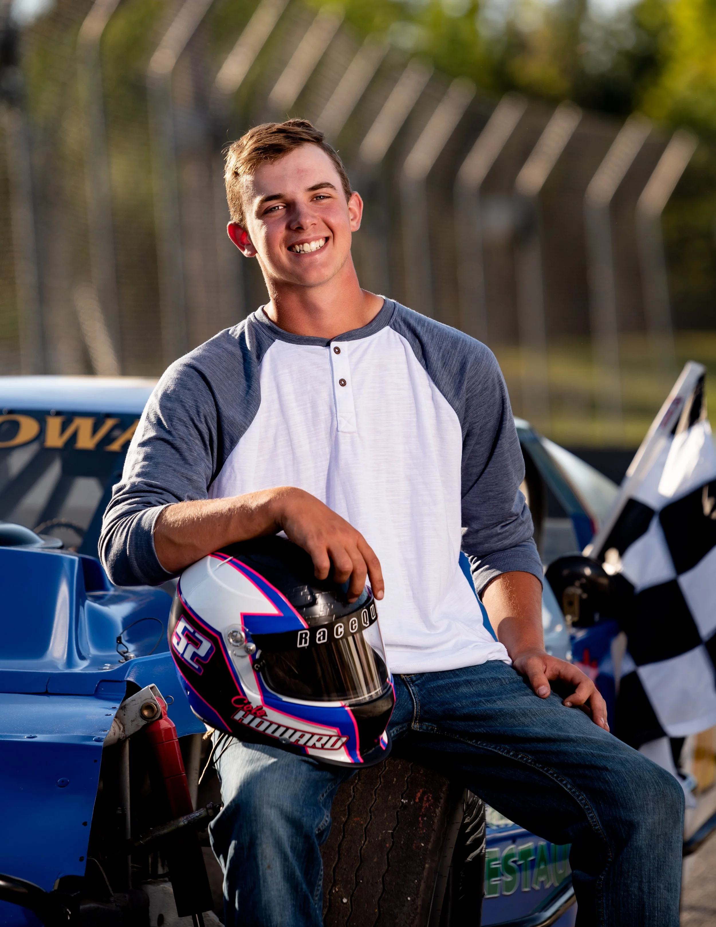 Young man holding a racing helmet, sitting on a tire next to a blue race car and a checkered flag, smiling during his race car inspired senior portraits in Wisconsin Dells.