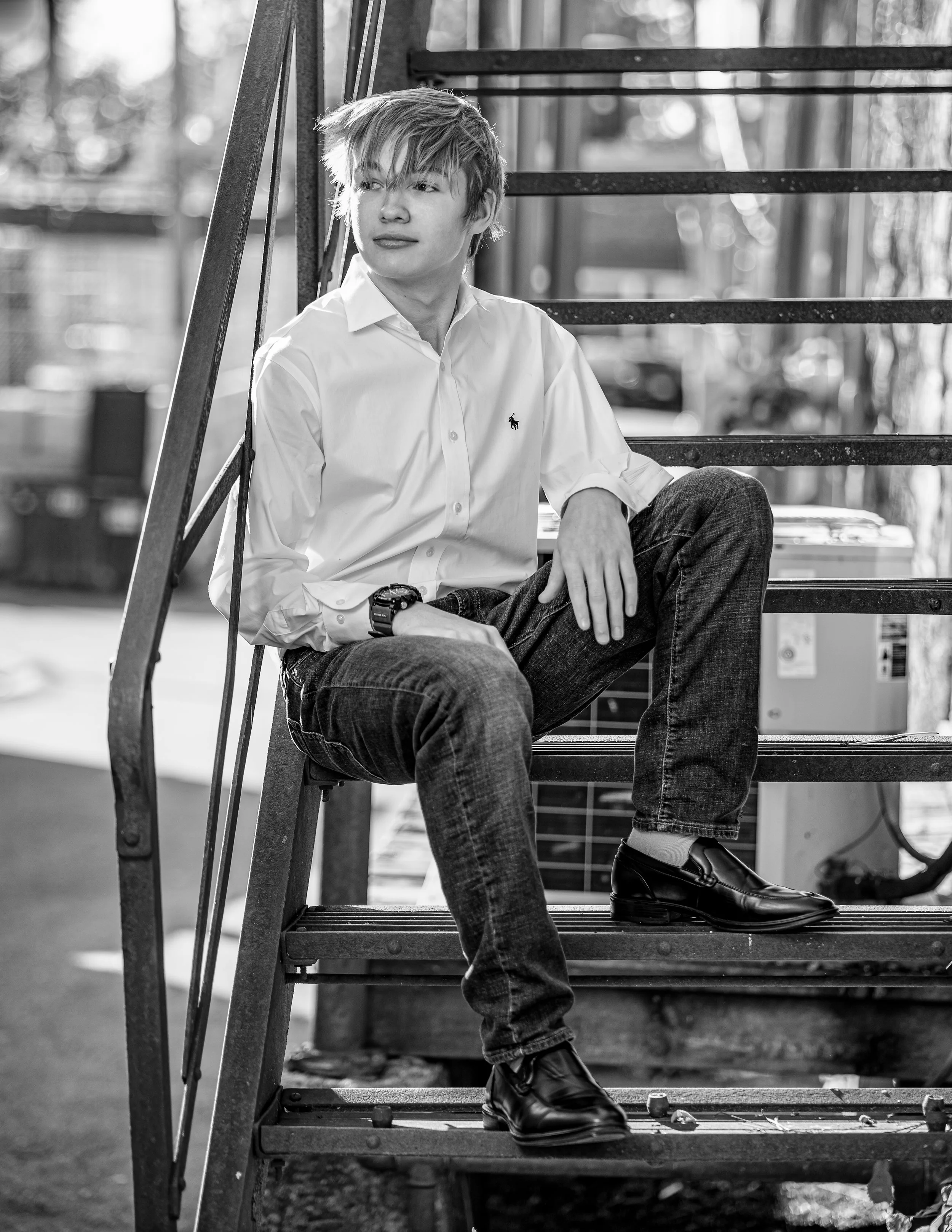 Black and white photo of a person sitting on metal stairs, wearing a white shirt and jeans, looking to the side.