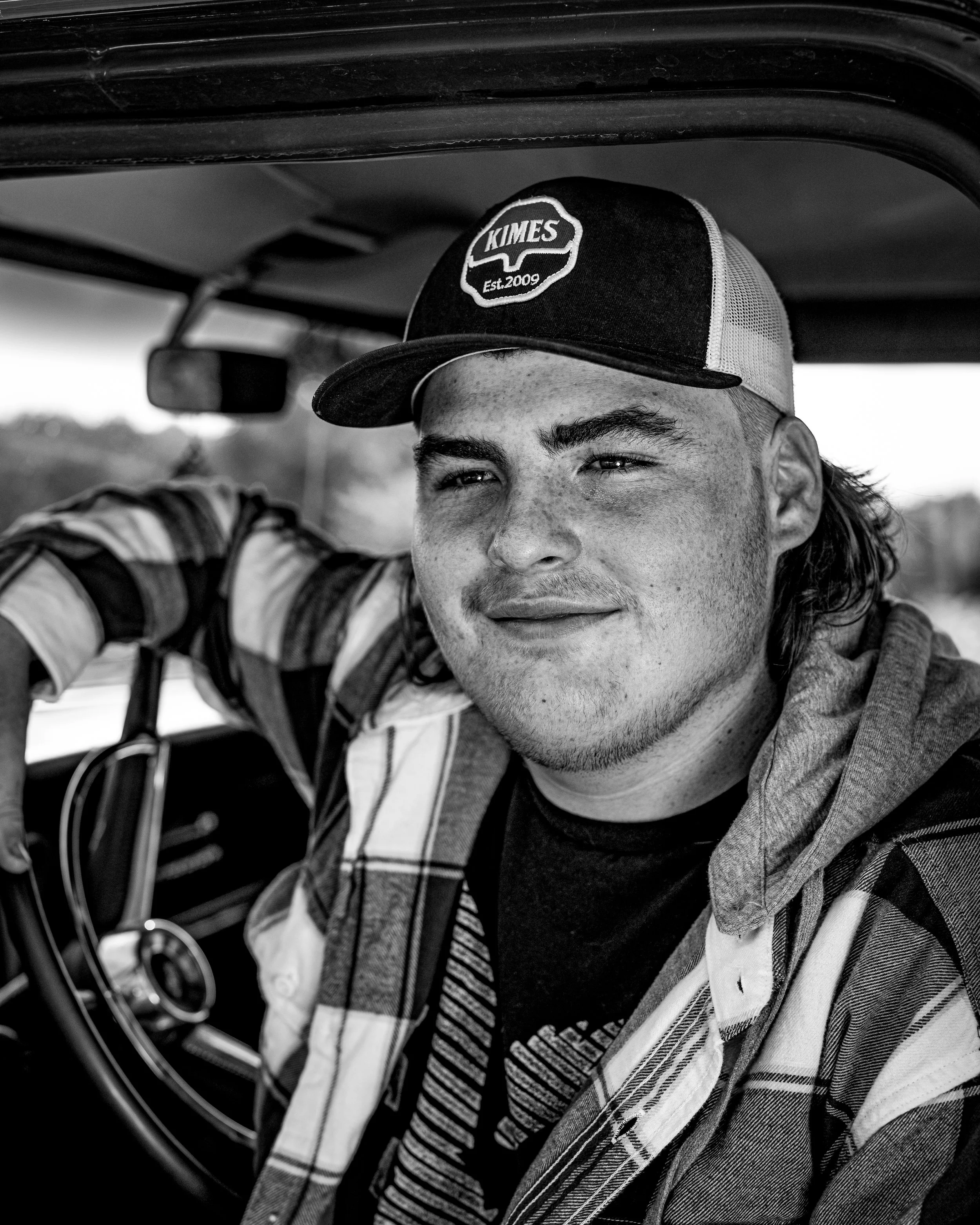Black and white portrait of a person in a flannel shirt and baseball cap sitting in a vehicle.