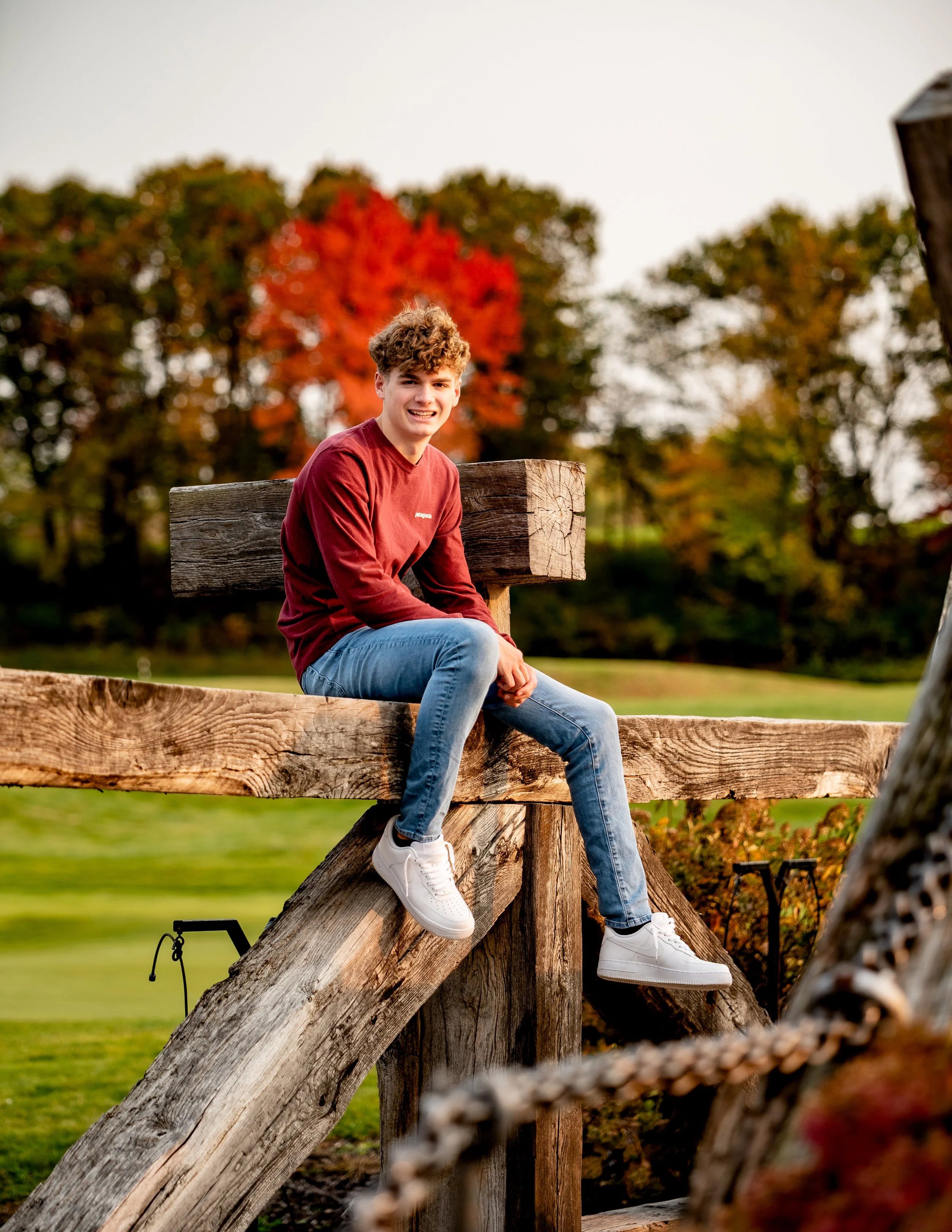 Person in a maroon long-sleeve shirt and jeans sitting on a wooden structure outdoors with fall foliage in the background.