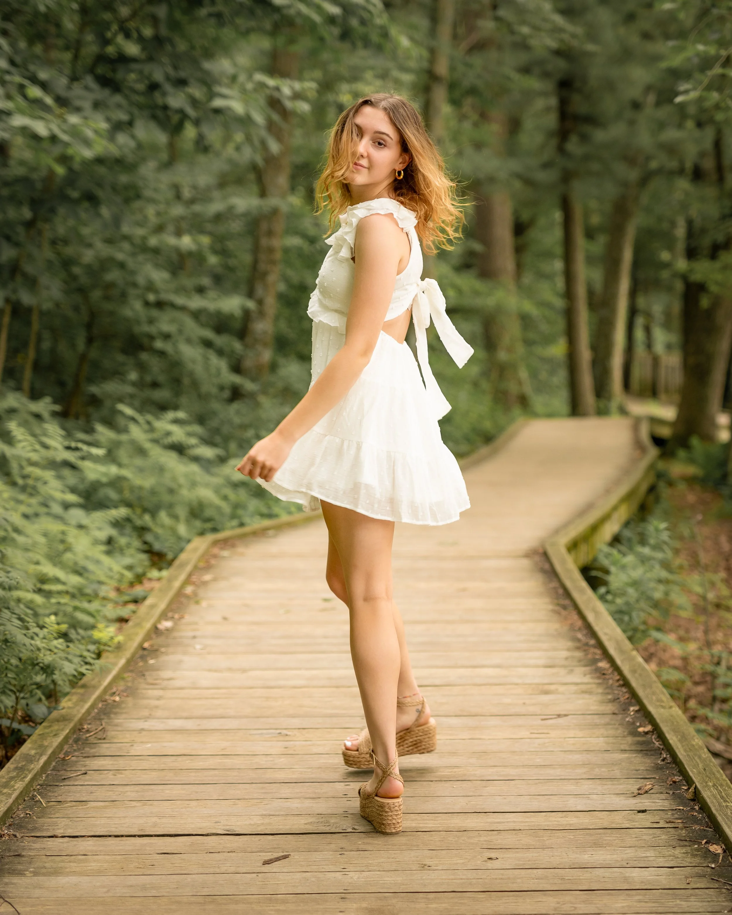 A woman in a white dress and wedge sandals walking on a wooden boardwalk through a lush green forest.