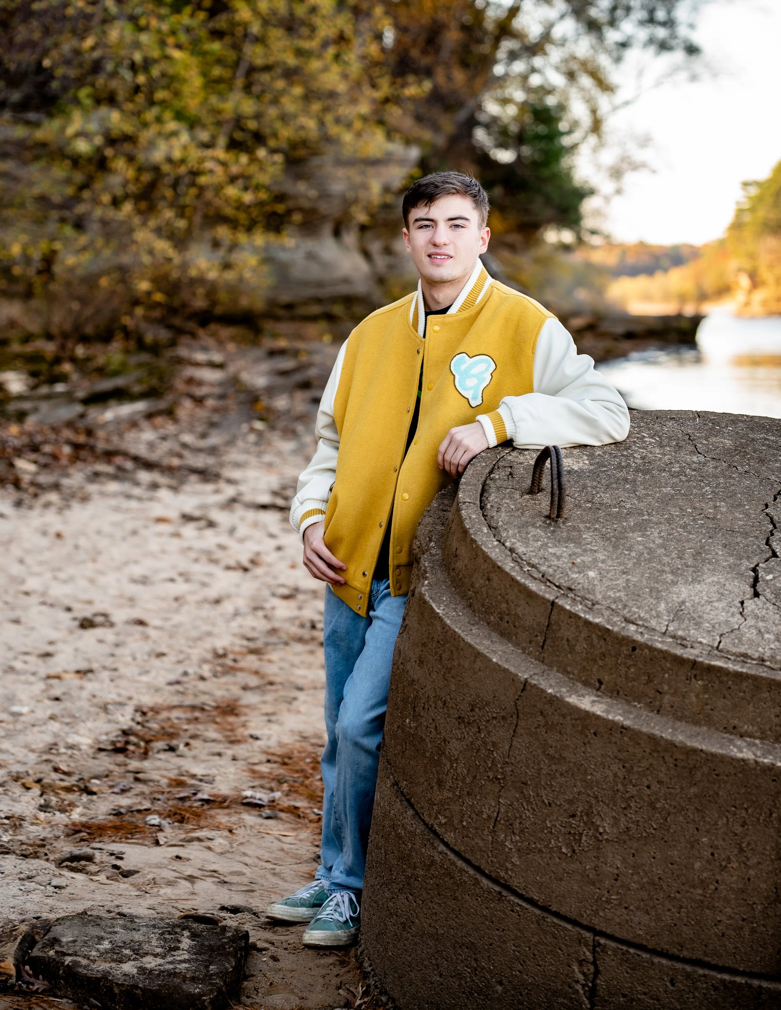 A young man in a yellow varsity jacket leans against a large concrete structure by a riverbank. He is surrounded by autumn foliage and wears light blue jeans and sneakers.
