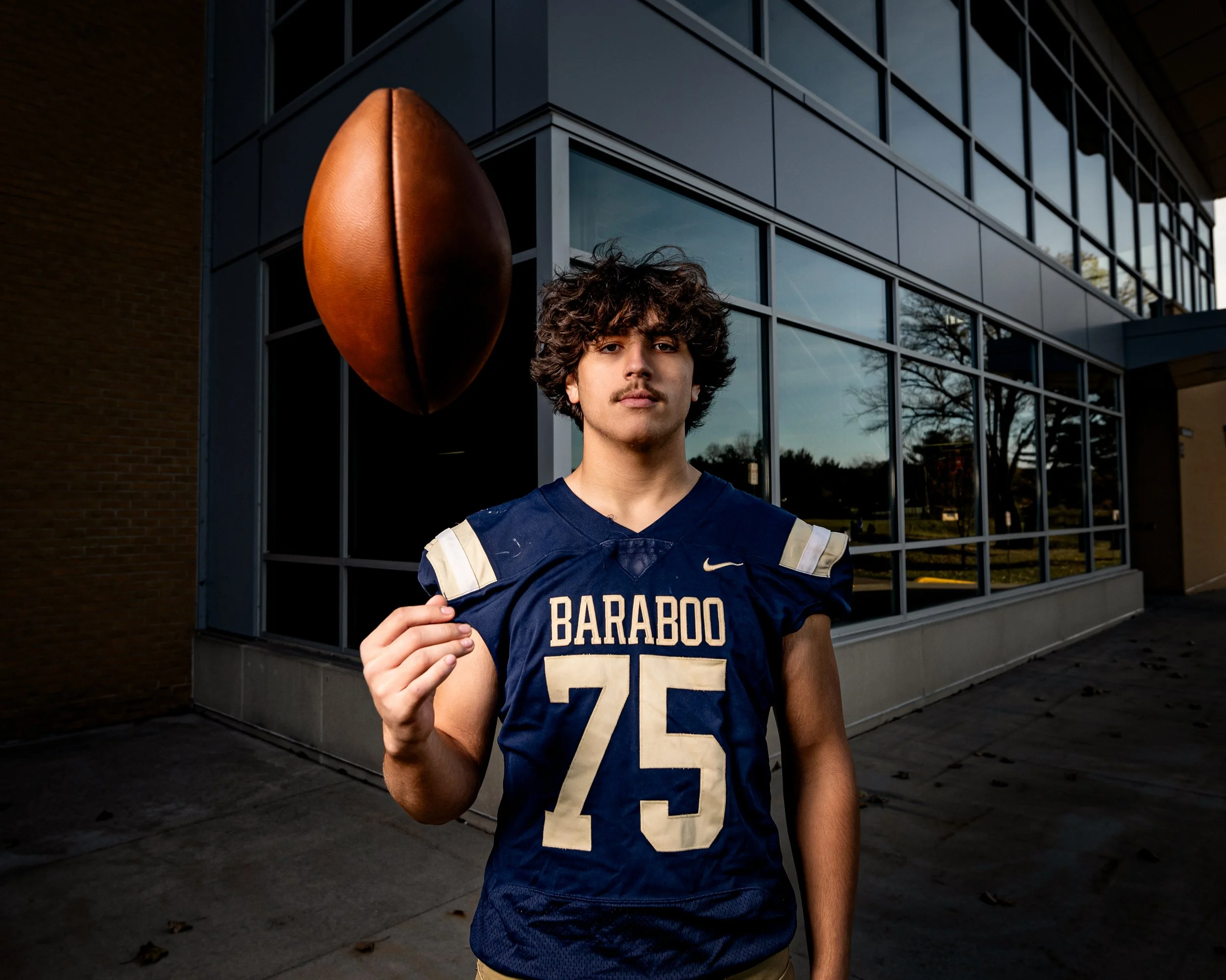 A young man wearing "Baraboo" football jersey with number 75 is spinning a football on their index finger outside a building with large window for his senior portraits.