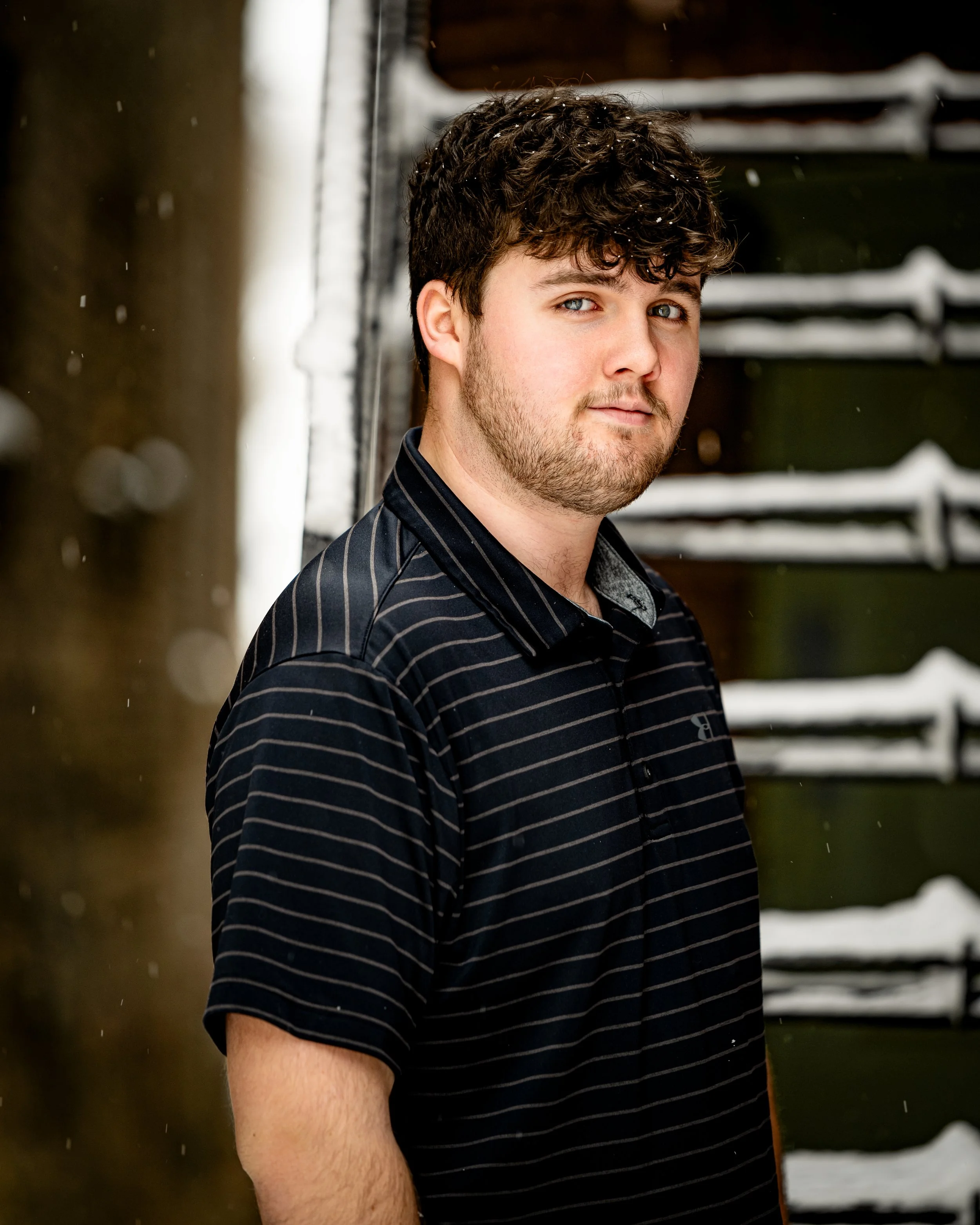 Man in black striped polo shirt standing in front of snow-covered railings during his senior portrait session in Baraboo,WI.