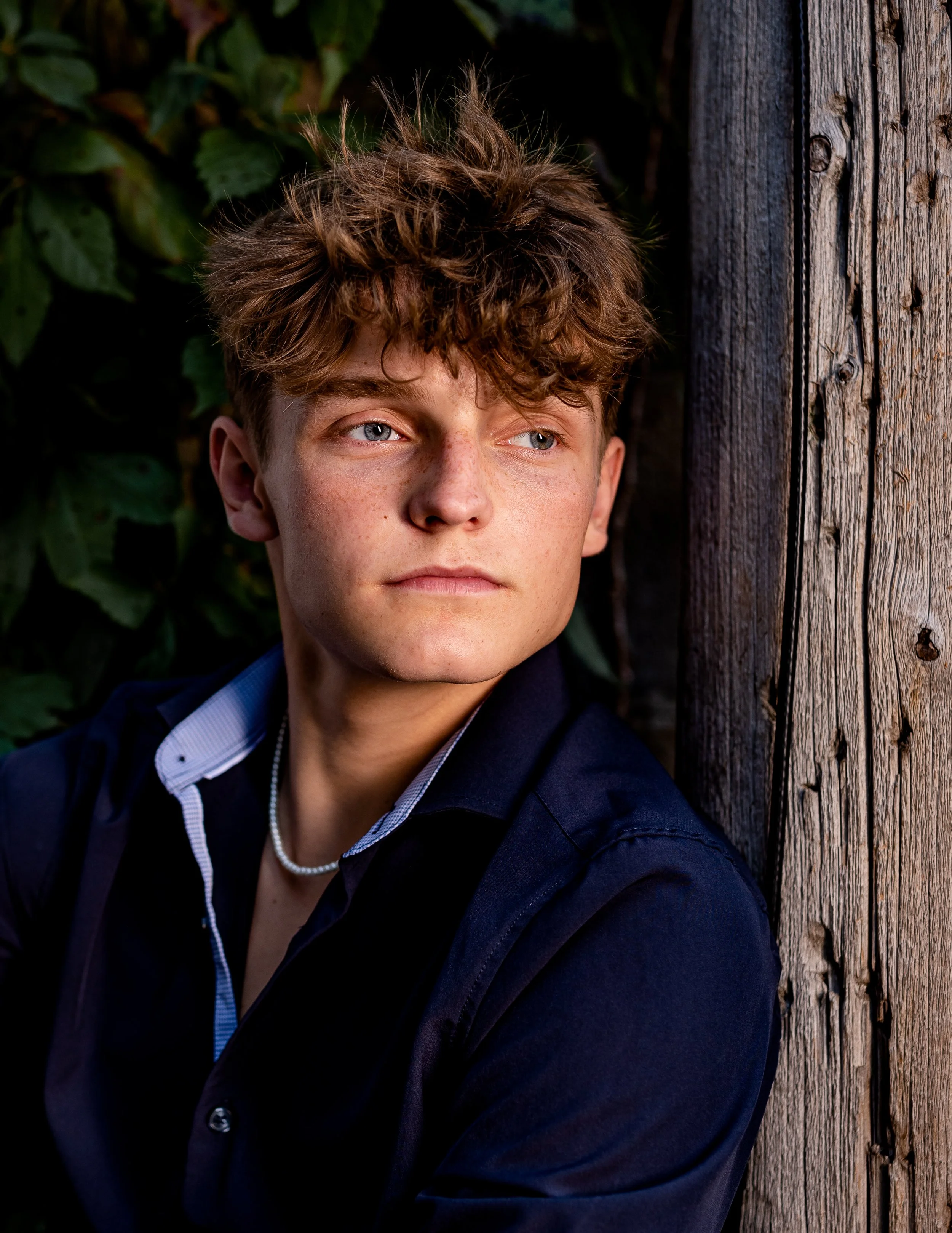 Young man with curly hair wearing a dark shirt and necklace, standing outdoors near a wooden structure and foliage.