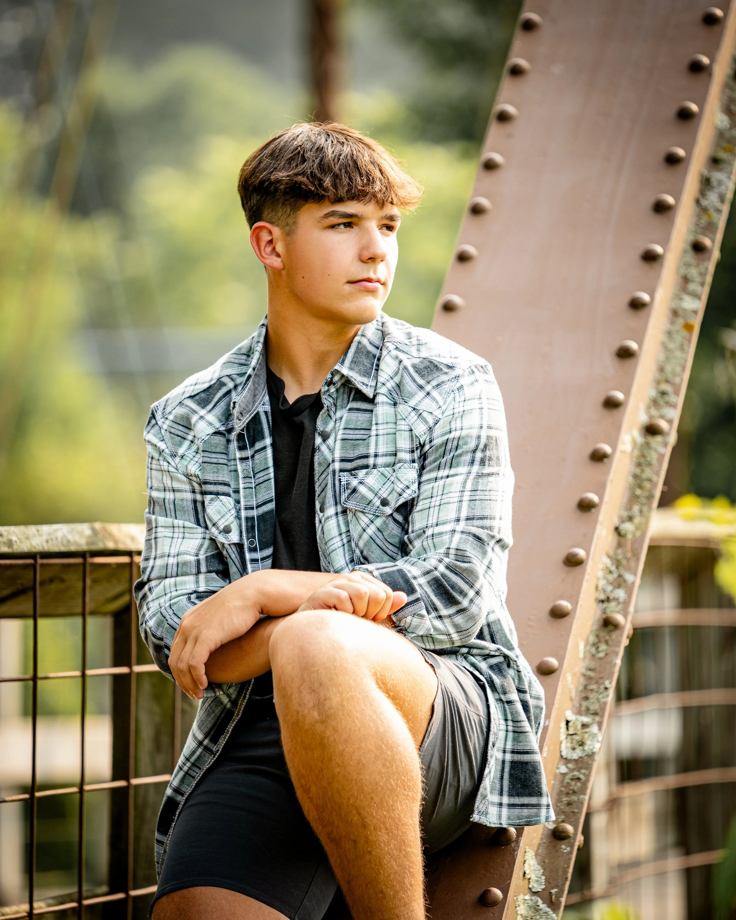 Young man sitting on a bridge, wearing a plaid shirt and shorts, looking thoughtfully to the side.