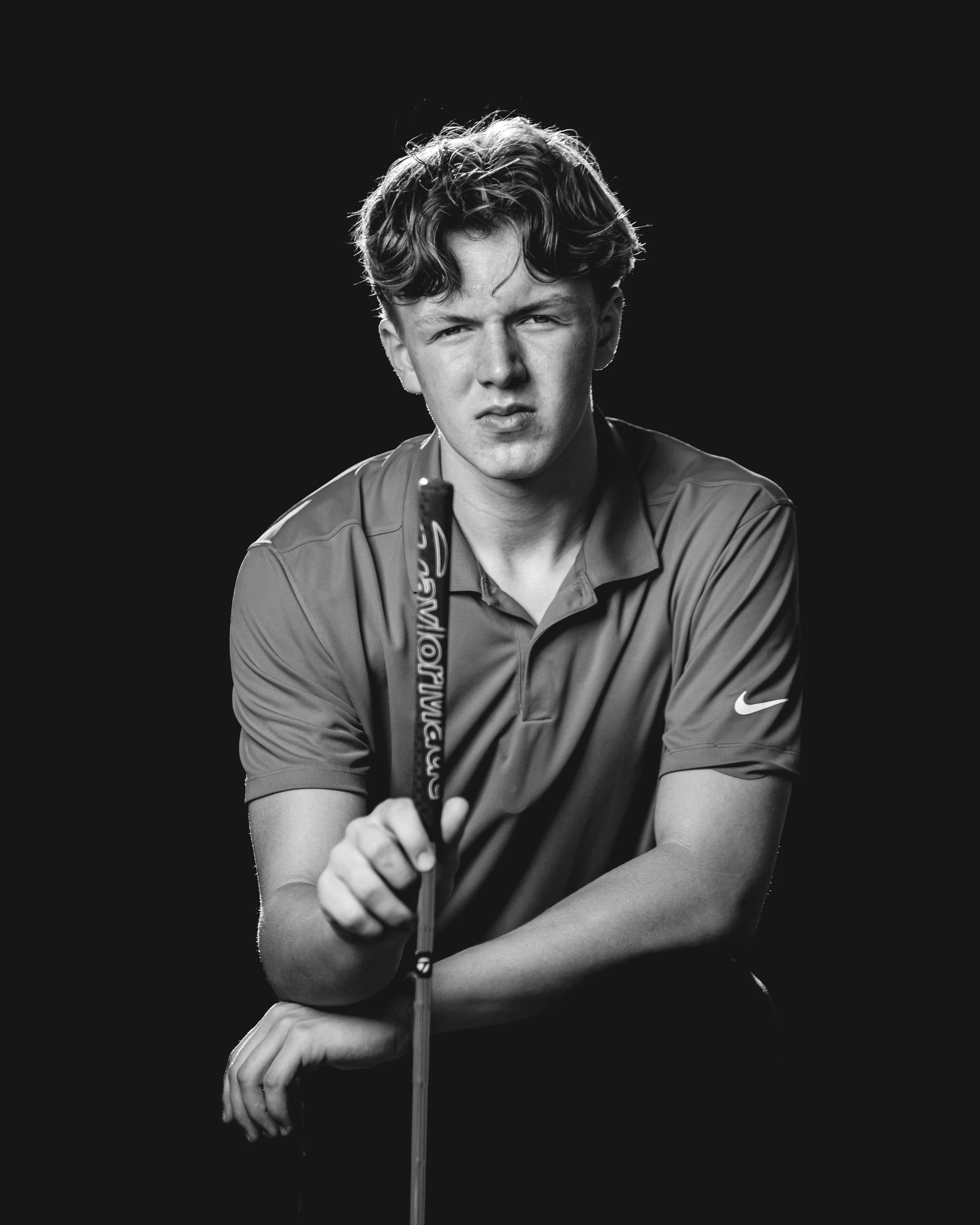 Black and white portrait of a young person holding a golf club, wearing a polo shirt with a Nike logo for his Tiger Woods inspired senior photos in Wisconsin Dells.