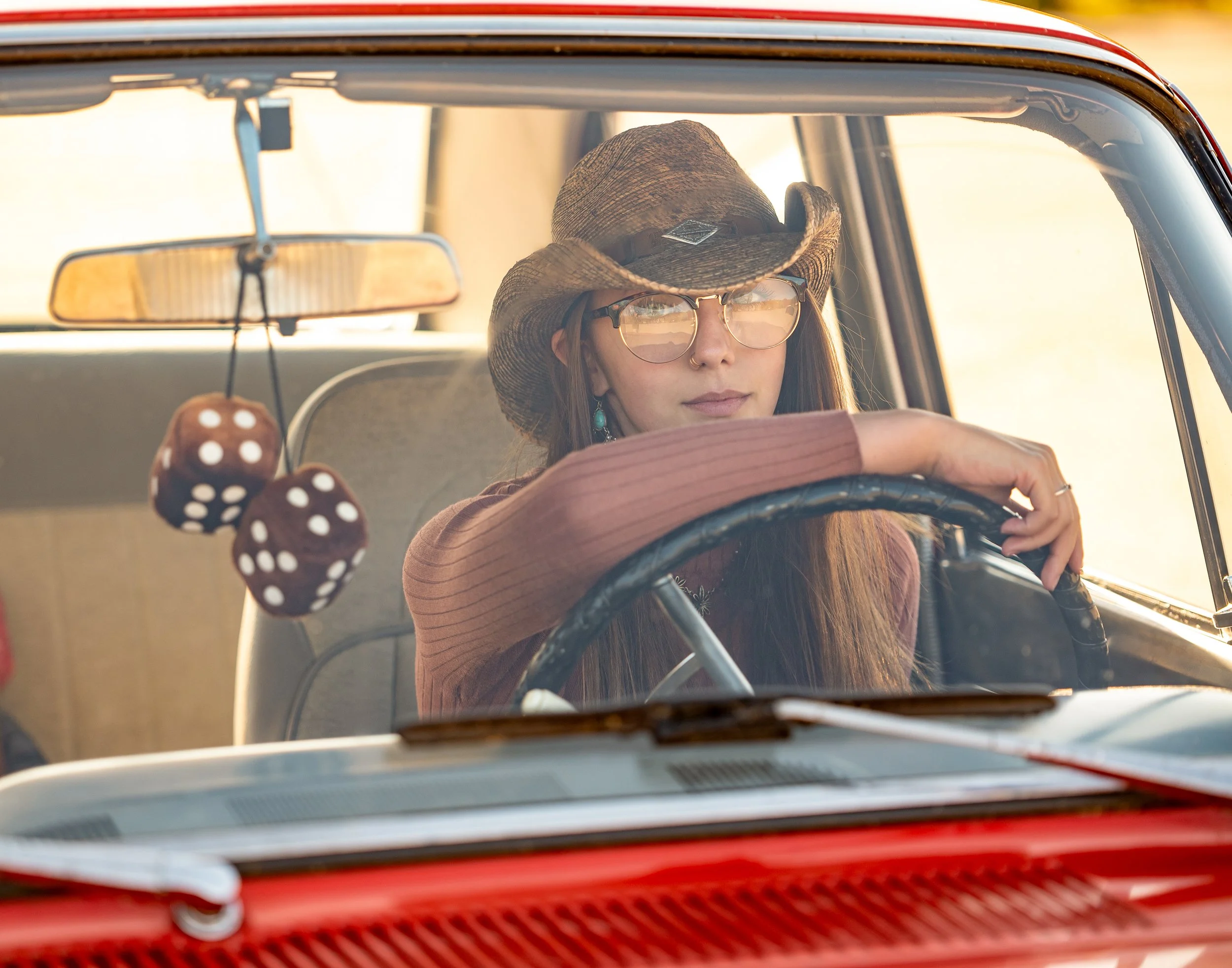 A woman wearing a cowboy hat and sunglasses driving a vintage red car with fuzzy dice hanging from the rearview mirror.