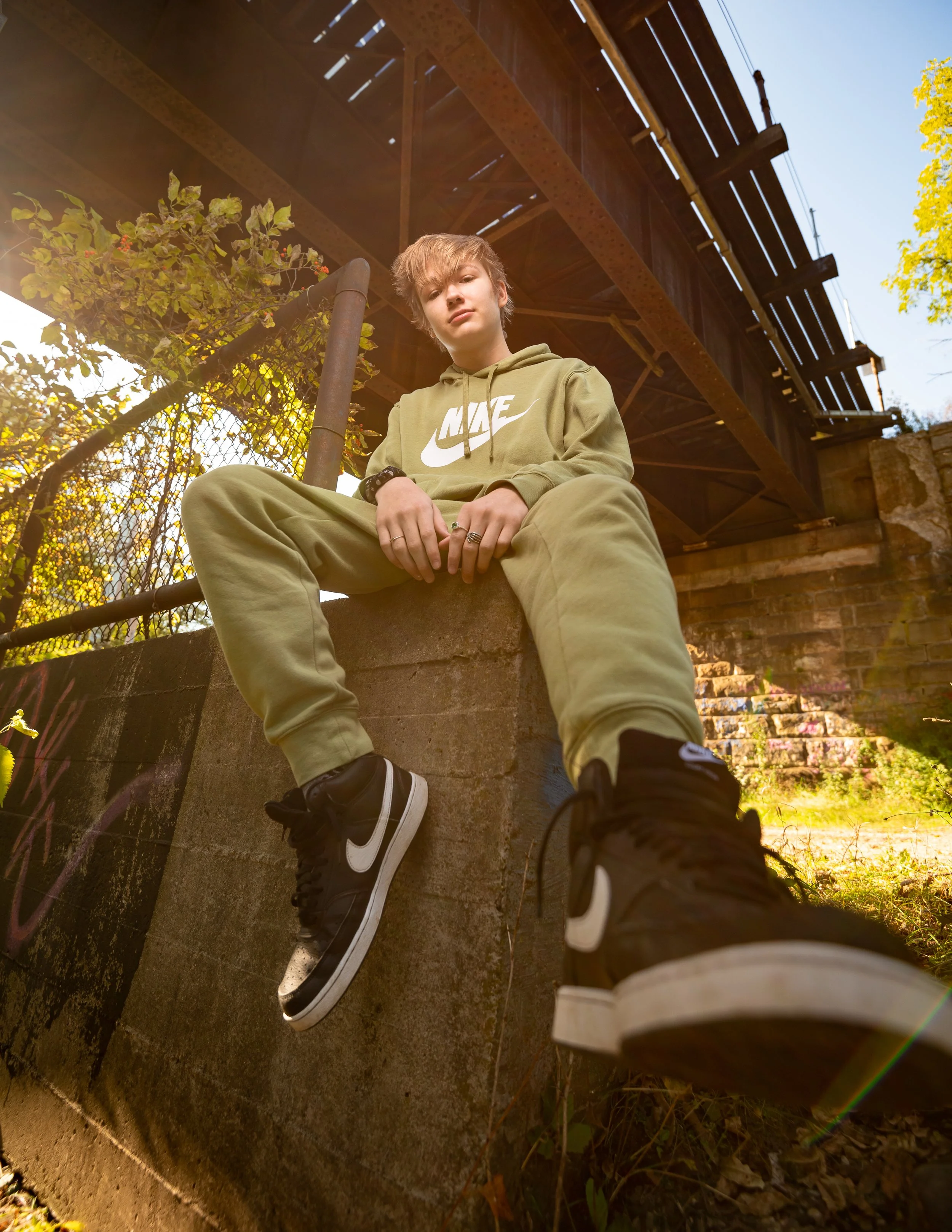 A person in a green Nike tracksuit sitting on a concrete ledge under a bridge, with sunlight streaming through. They are wearing black and white Nike sneakers and have a relaxed pose.