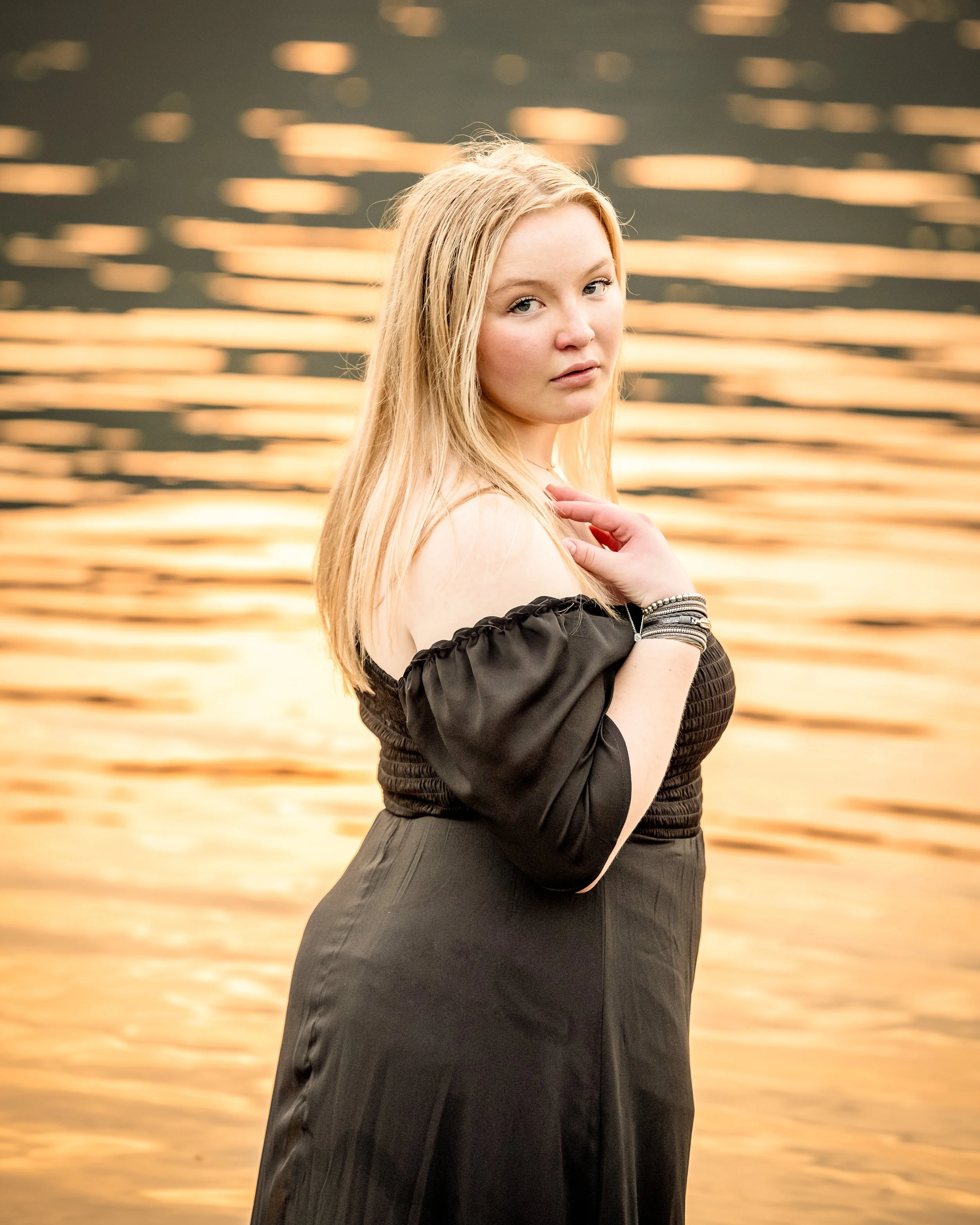 Woman in a black dress standing by water during sunset.