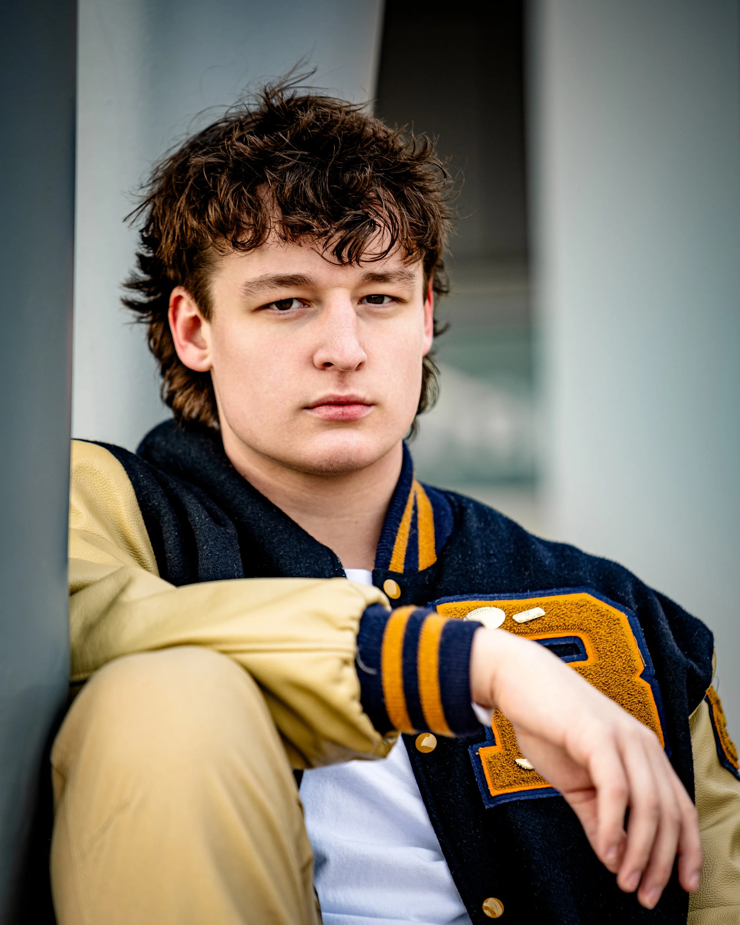 Young person wearing a varsity jacket with letter "B", sitting against a wall with a thoughtful expression.