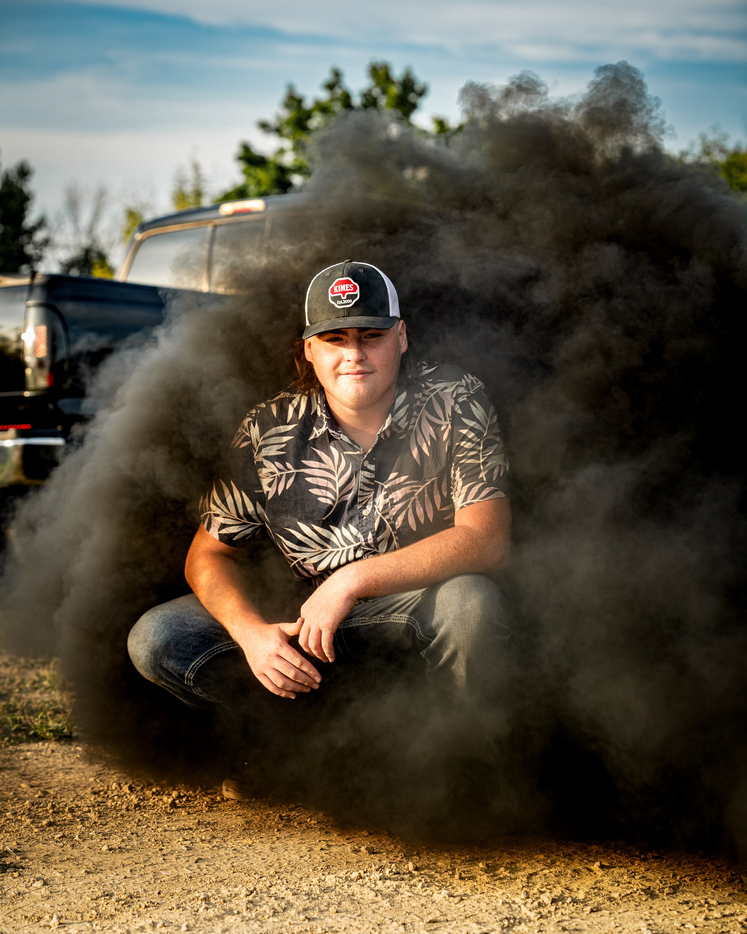 A person in a tropical shirt and cap crouching near a truck with thick black smoke around them, outdoors.