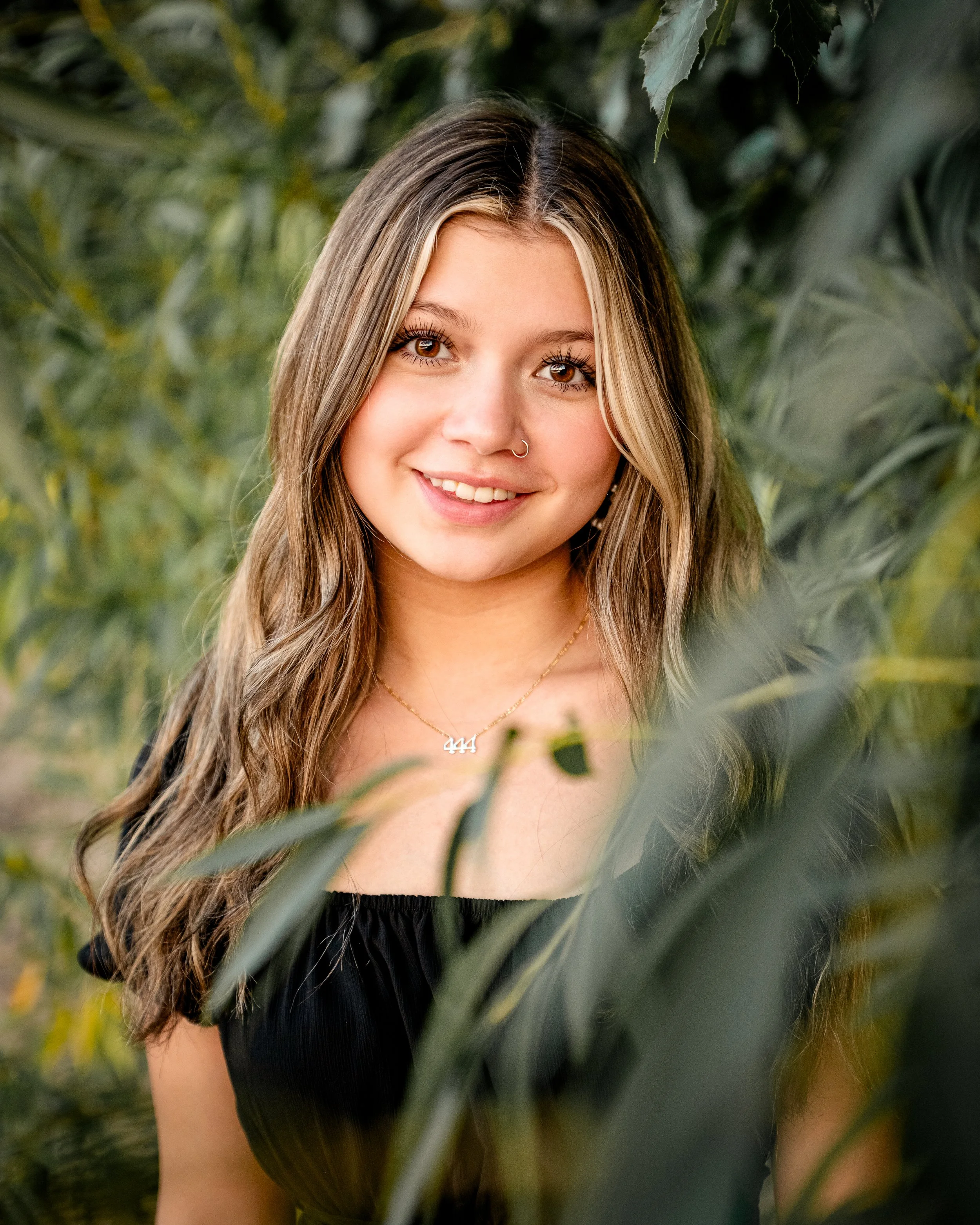 Woman with long hair and nose piercing smiling, surrounded by green foliage.