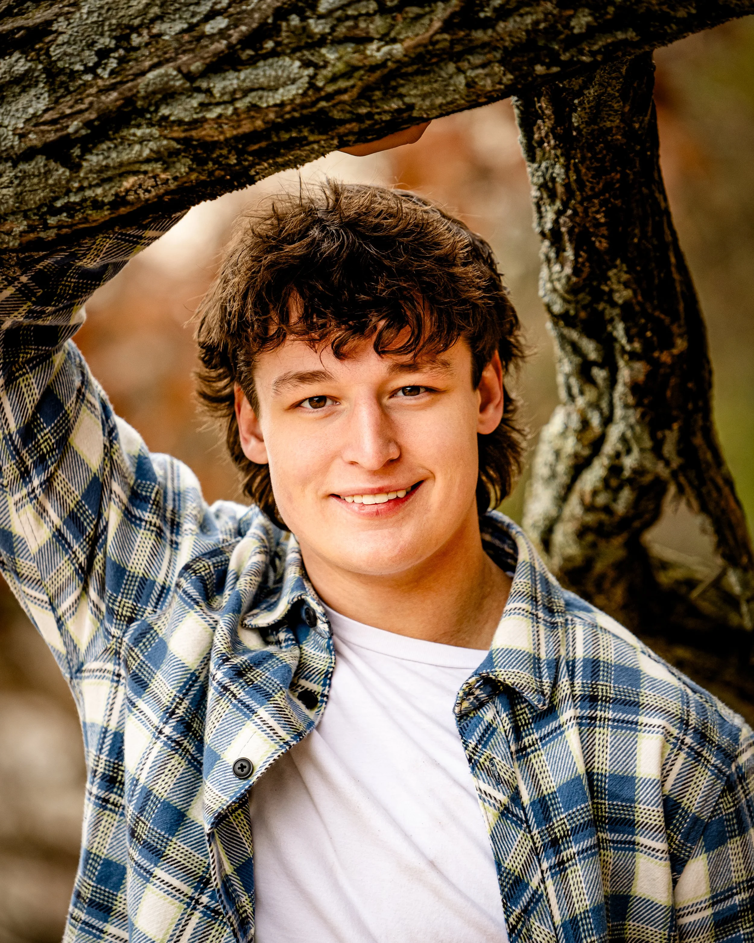 A young man smiling in plaid shirt leaning against a tree branch outdoors for his senior photos near Wisconsin Dells.