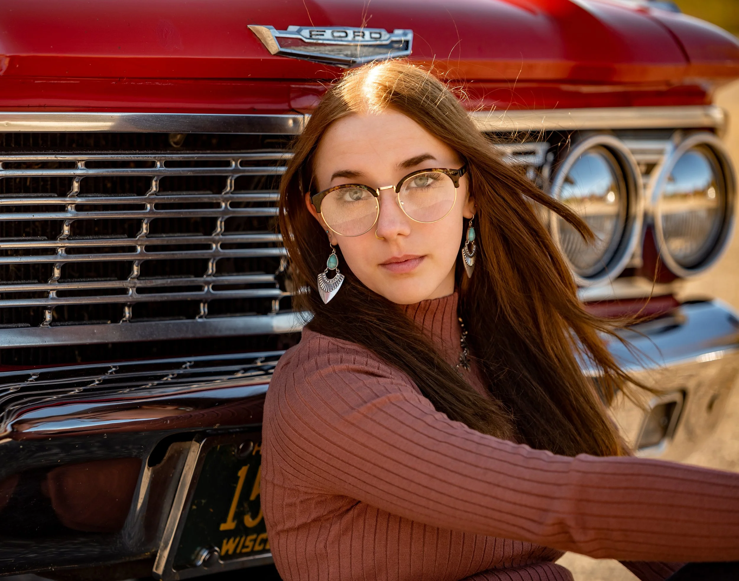 Young person with glasses and long brown hair sitting in front of a vintage red Ford car, wearing a brown ribbed sweater and decorative earrings.