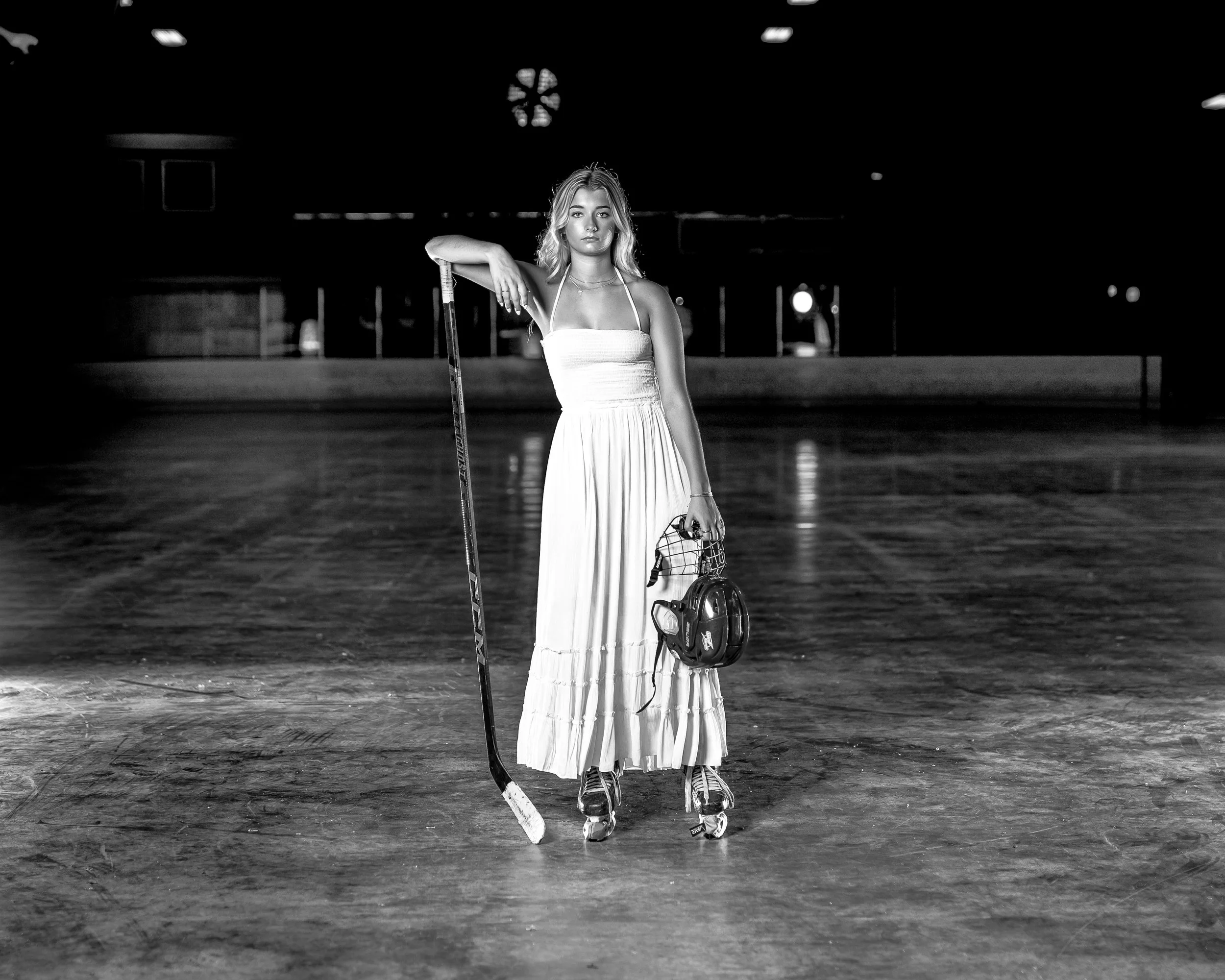 Young woman in a dress on an ice rink holding a hockey stick and a helmet, wearing ice skates for a dramatic black and white senior picture in Baraboo.
