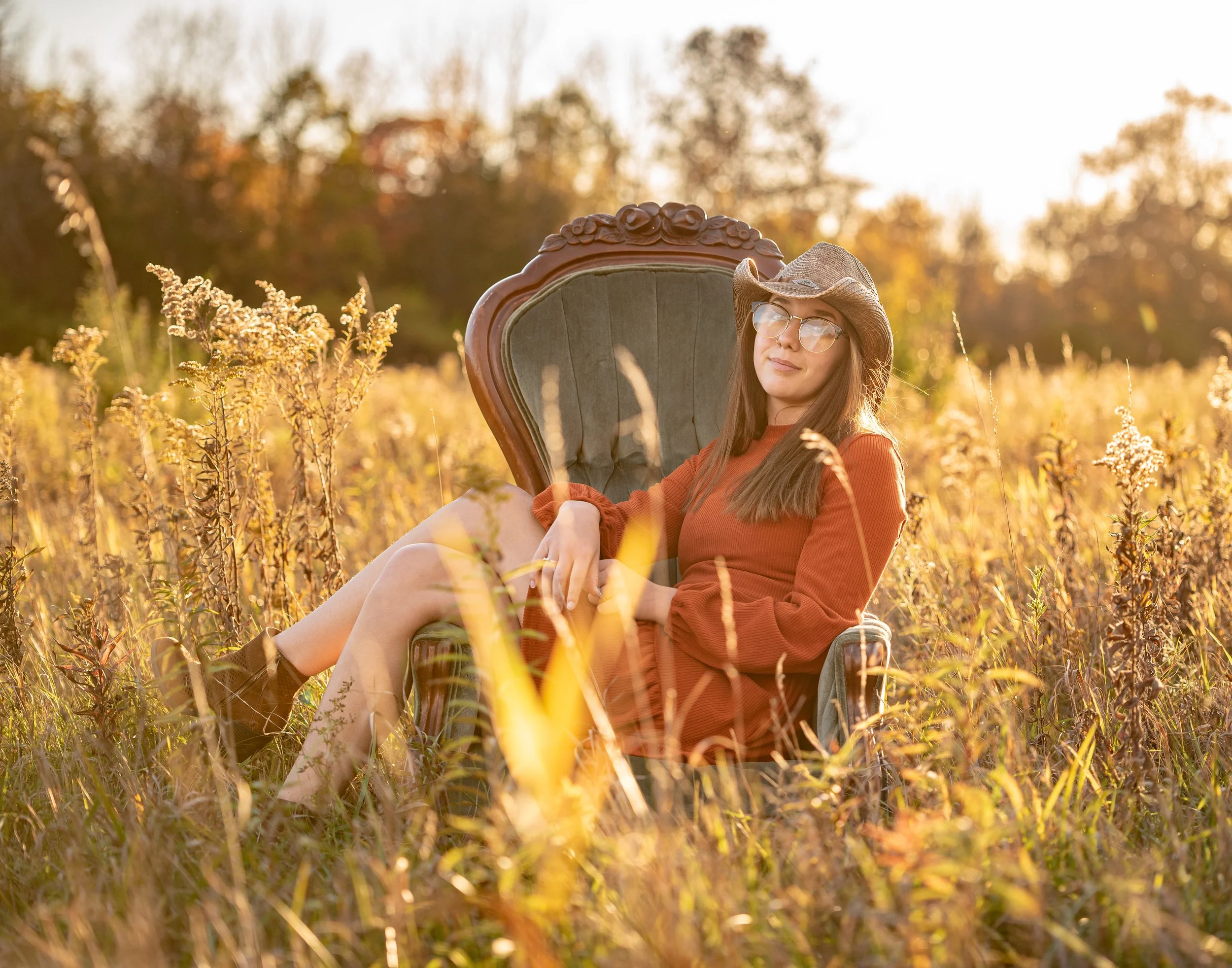 A woman in a hat sitting on a vintage chair in a sunlit field, surrounded by tall grass and wildflowers, during a golden hour scene in autumn.