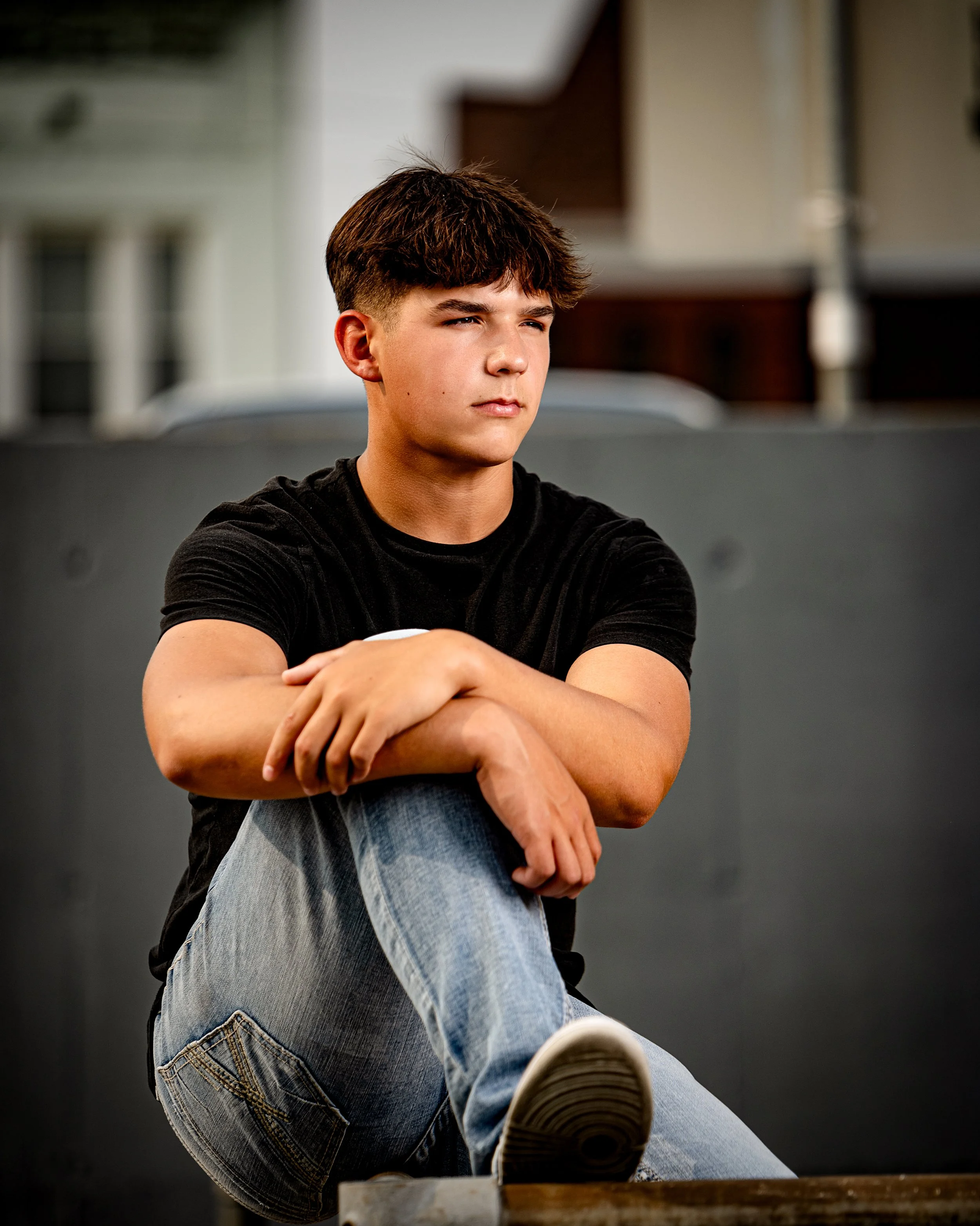 Young man sitting with arms crossed over knees, wearing a black t-shirt and jeans, against an urban background.