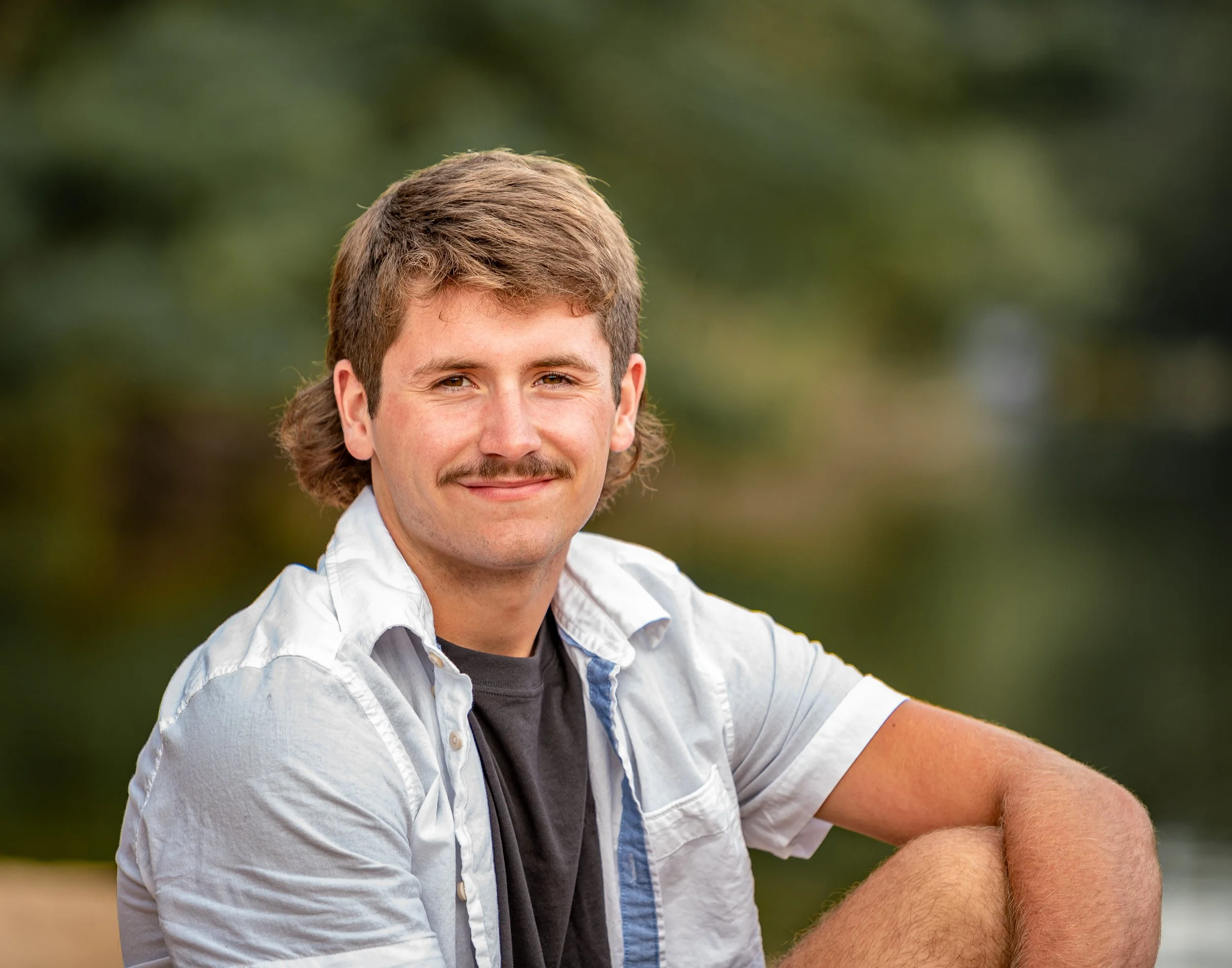 Man with a mustache and short brown hair sitting outdoors, wearing a white shirt over a black t-shirt, smiling at the camera. Background is blurred greenery.