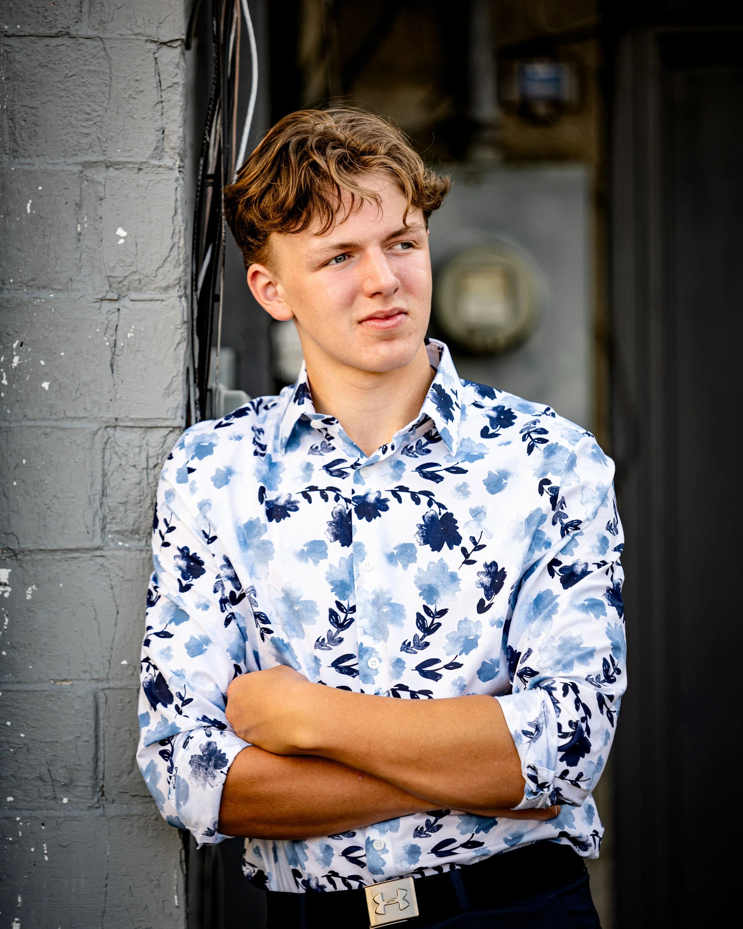 Young man in a floral shirt leaning against a gray wall, looking to the side with arms crossed in downtown Wisconsin Dells for senior portraits. 