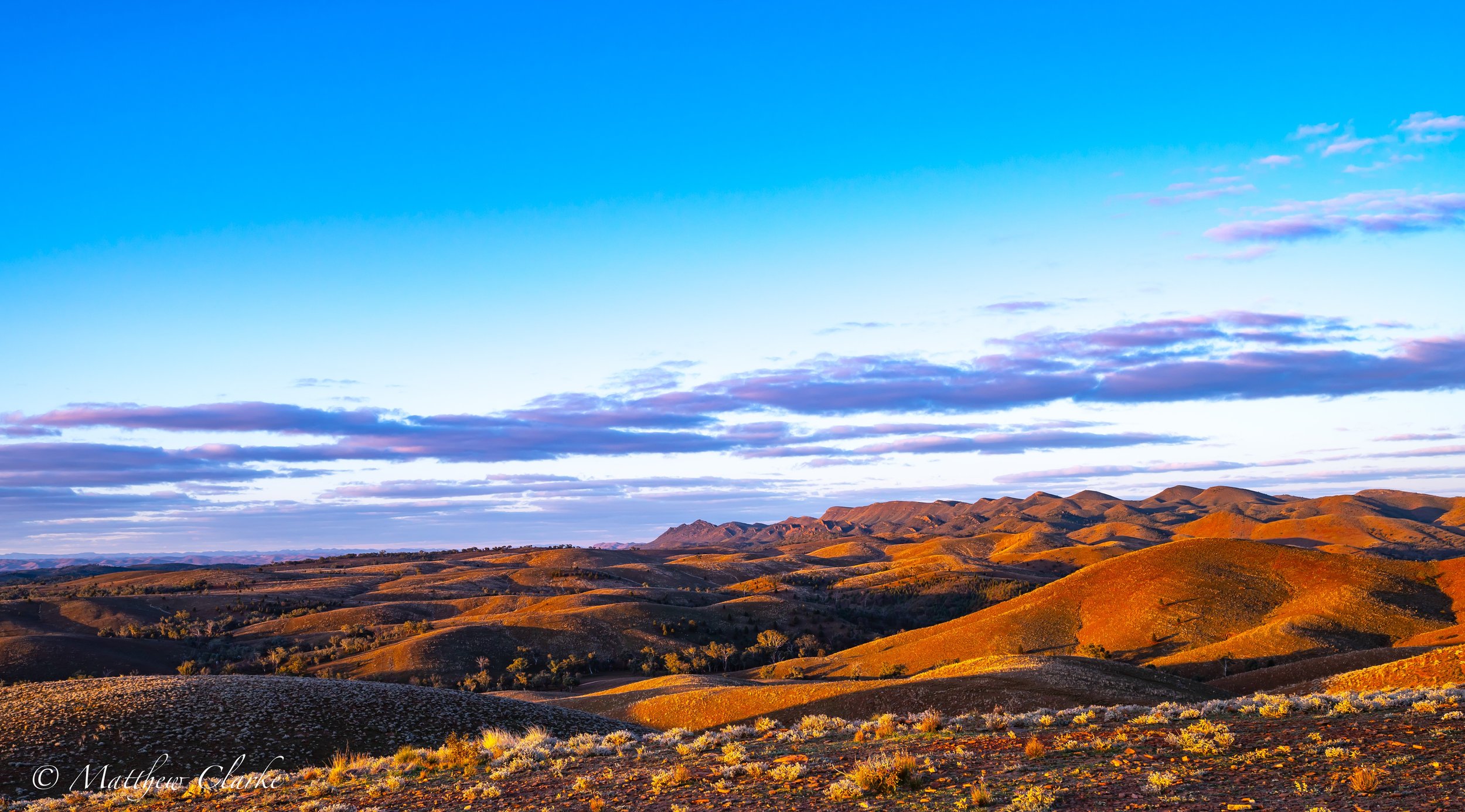 Rolling hills in the late afternoon with dry grass and sparse vegetation, under a sky with scattered clouds at sunset.