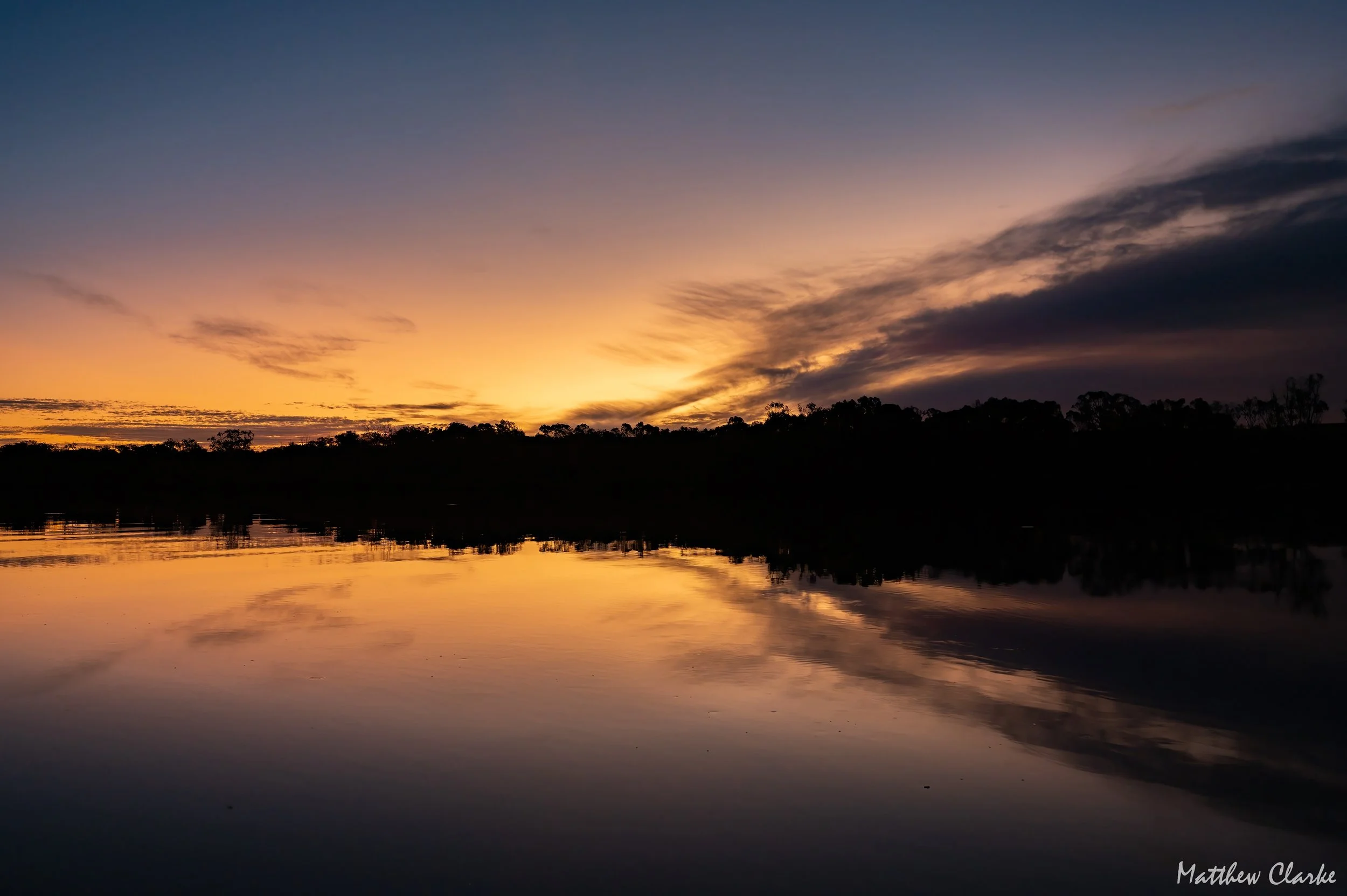 A peaceful sunset over a body of water with a silhouette of trees in the background and a reflection of the colorful sky on the water's surface.