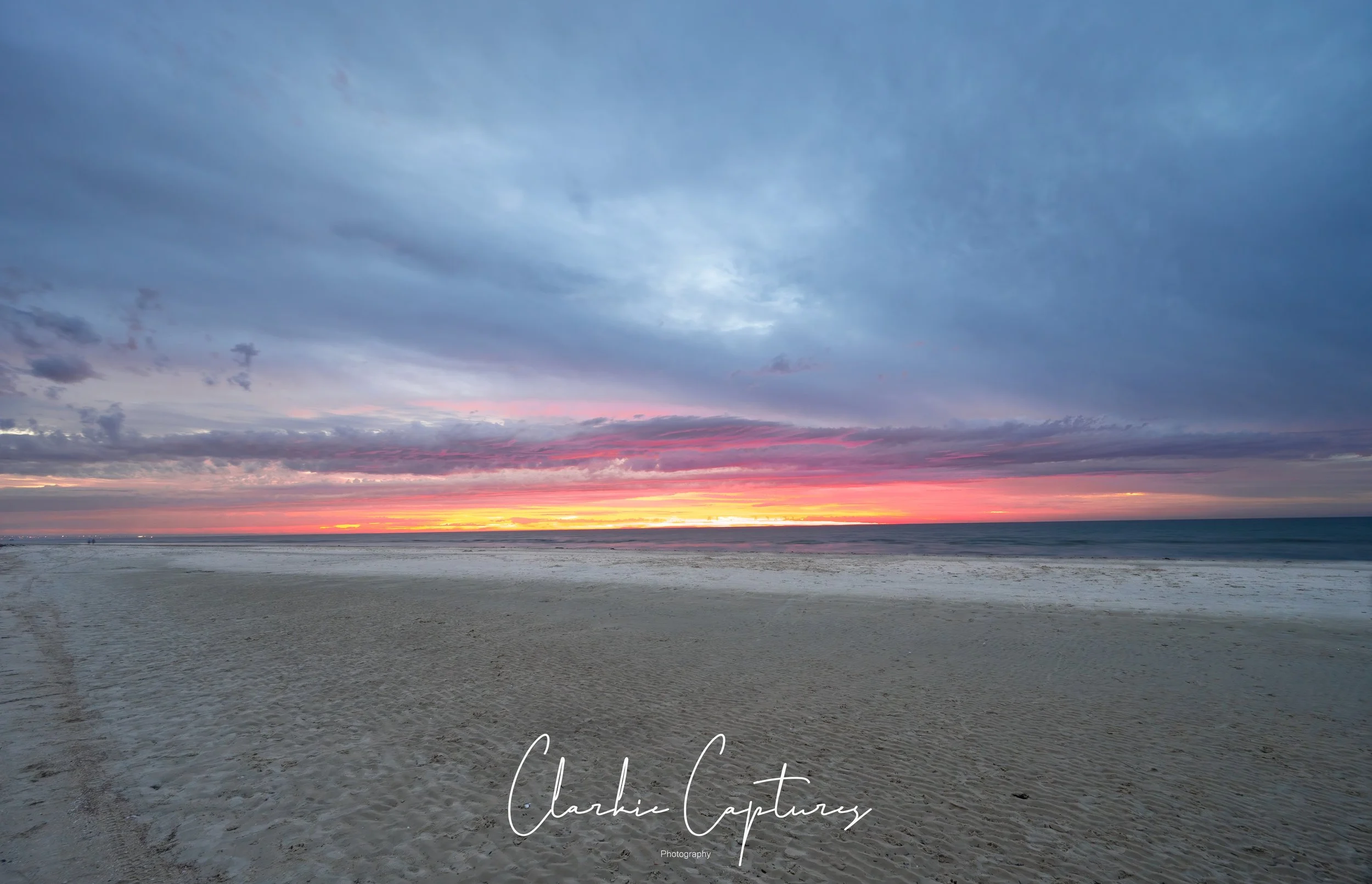 A beach scene at sunset with a colorful sky of pink, orange, and purple hues reflected on the ocean, sandy shore, and dramatic clouds.