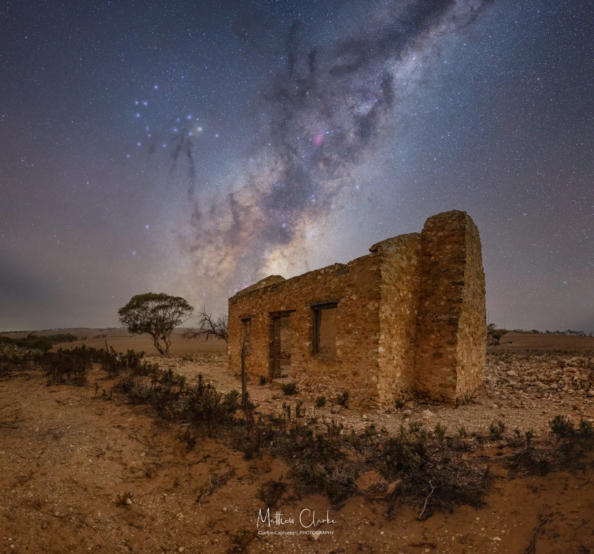 Night sky filled with stars and the Milky Way galaxy, with a silhouette of a building made of stone and a few trees in the foreground.