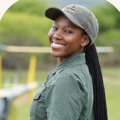 A woman smiling outdoors in a green uniform and baseball cap, with long braided hair, standing in a grassy area.