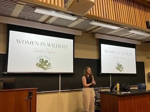 A woman giving a presentation in a classroom with two large screens displaying a slide titled "Women in Wildlife".