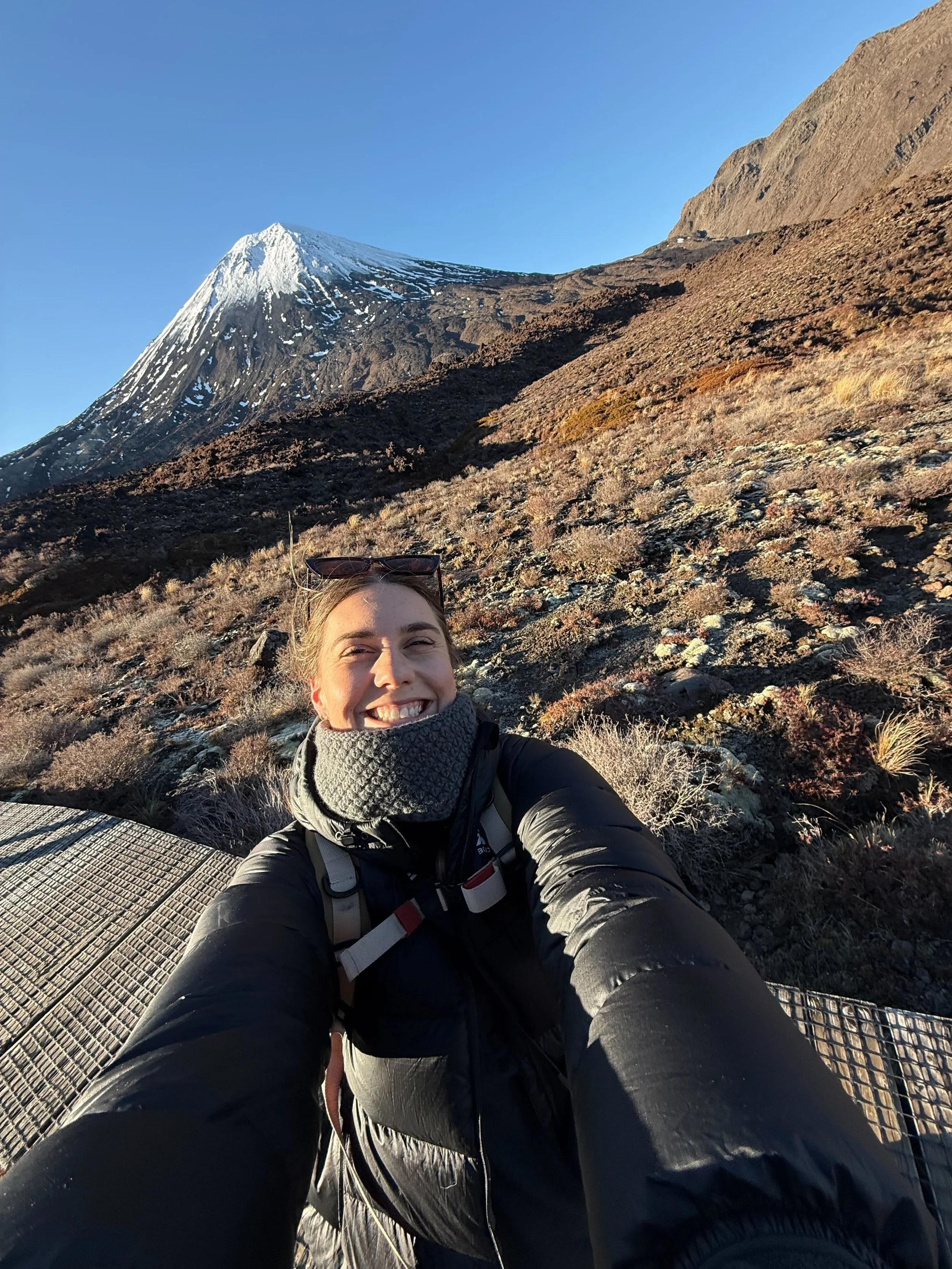A woman taking a selfie outdoors on a mountain trail with a snow-capped mountain in the background, wearing a black jacket, gray scarf, and sunglasses.