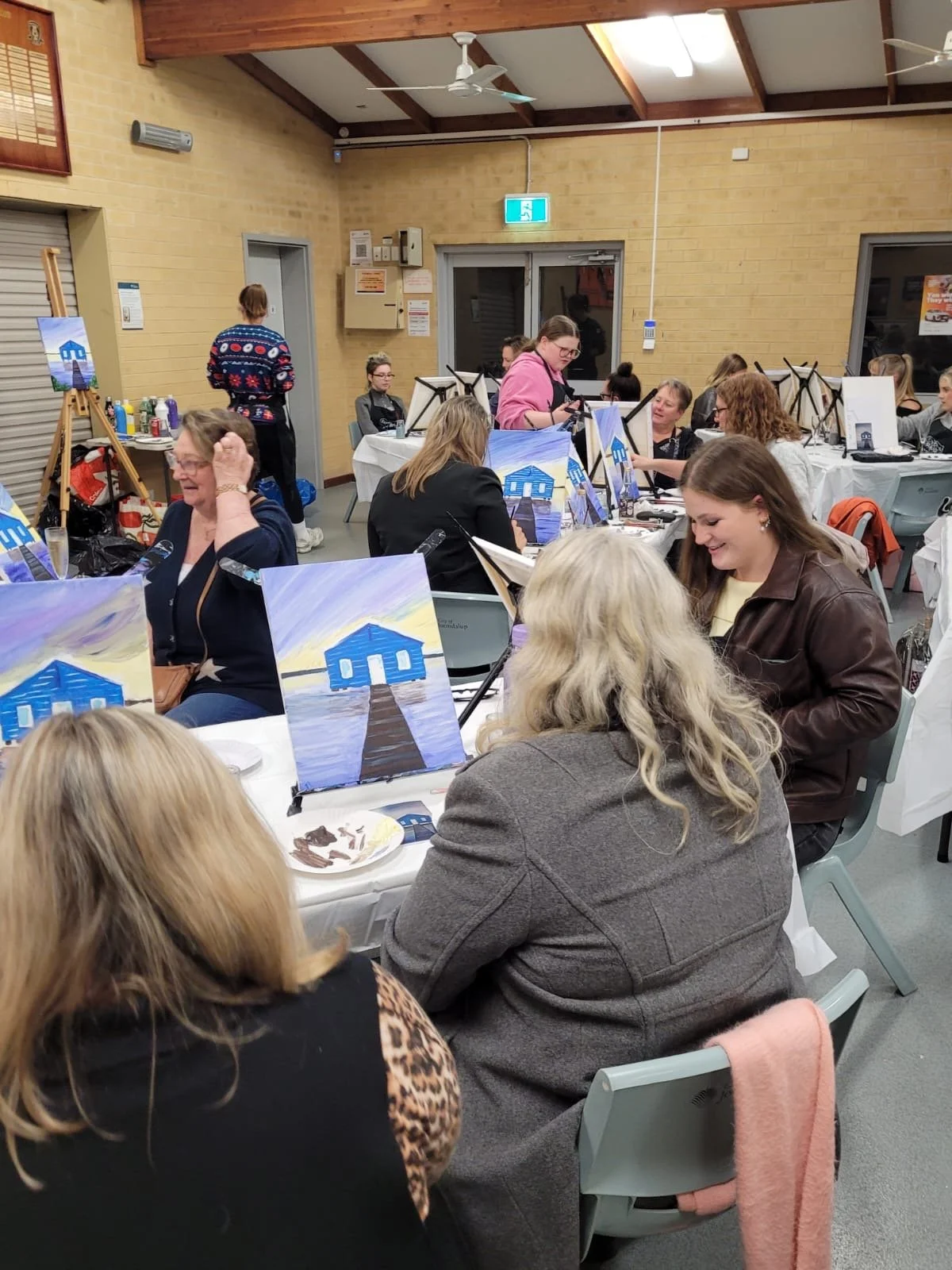 People participating in a painting class, painting pictures of a blue house on canvases. The room has beige brick walls, a wooden ceiling, and large windows. Participants are seated at tables covered with white tablecloths, and there are painting supplies and finished paintings visible.