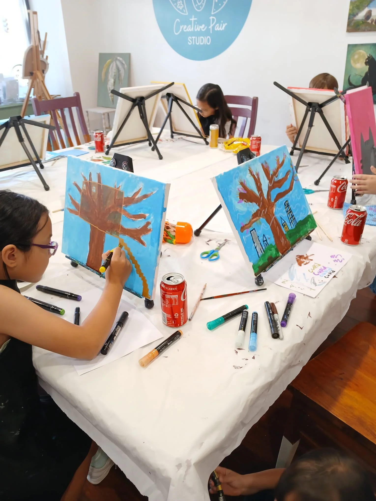 Children painting pictures of large trees on small canvases at a group art class in a studio.
