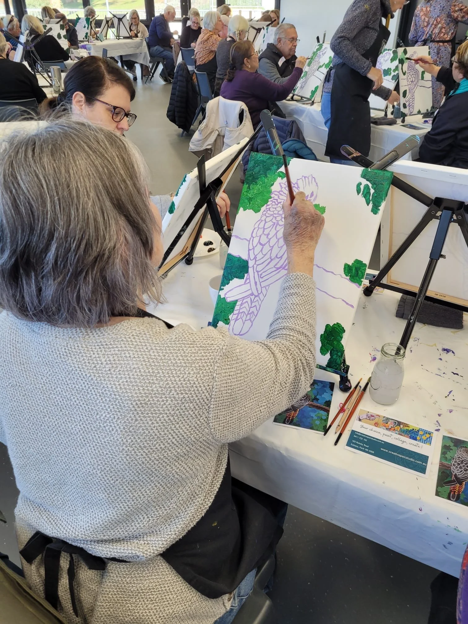 A group of people participating in a painting class, with some working on a landscape painting of a bird perched on a branch surrounded by green foliage, while others are in the background working on their own paintings.