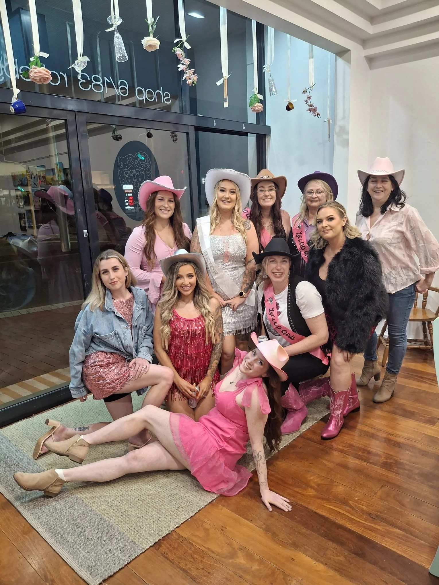 A group of eleven women dressed in cowboy and cowgirl attire, including hats and boots, posing inside a store with wooden floors and a glass storefront. Some women wear sashes that say "Je t'aime" and "Courage," and one woman is lying on the floor in a pink dress and cowboy hat.
