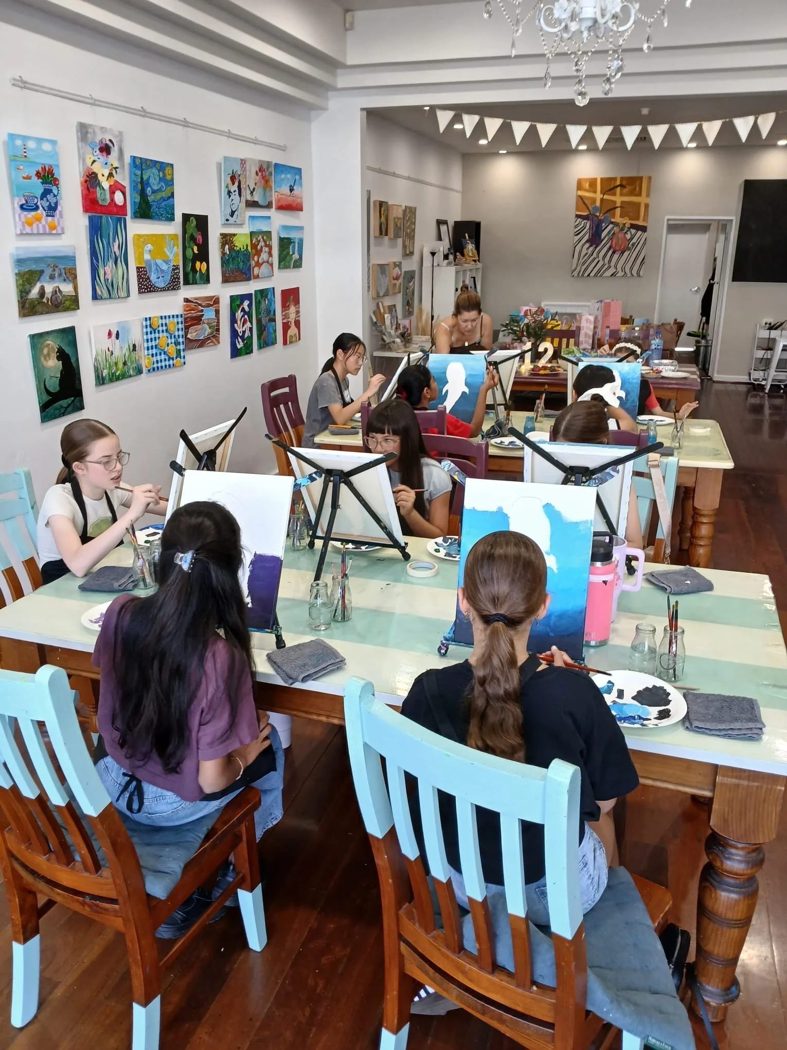 Children participating in a painting class in an art studio, with their canvases and art supplies on tables. The studio has colorful paintings on the walls and a chandelier hanging from the ceiling.