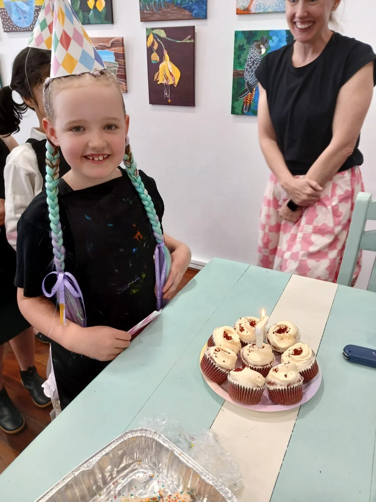 A young girl with colorful braided hair, wearing a birthday hat, stands next to a cupcake cake with one lit candle. An adult woman, smiling, stands behind her. The scene appears to be a birthday celebration at an art gallery, with paintings on the wall.