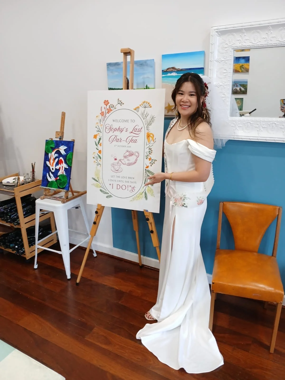 A woman in a white dress standing next to a signboard at an art gallery or studio, with paintings on the wall and a wooden floor, smiling at the camera.