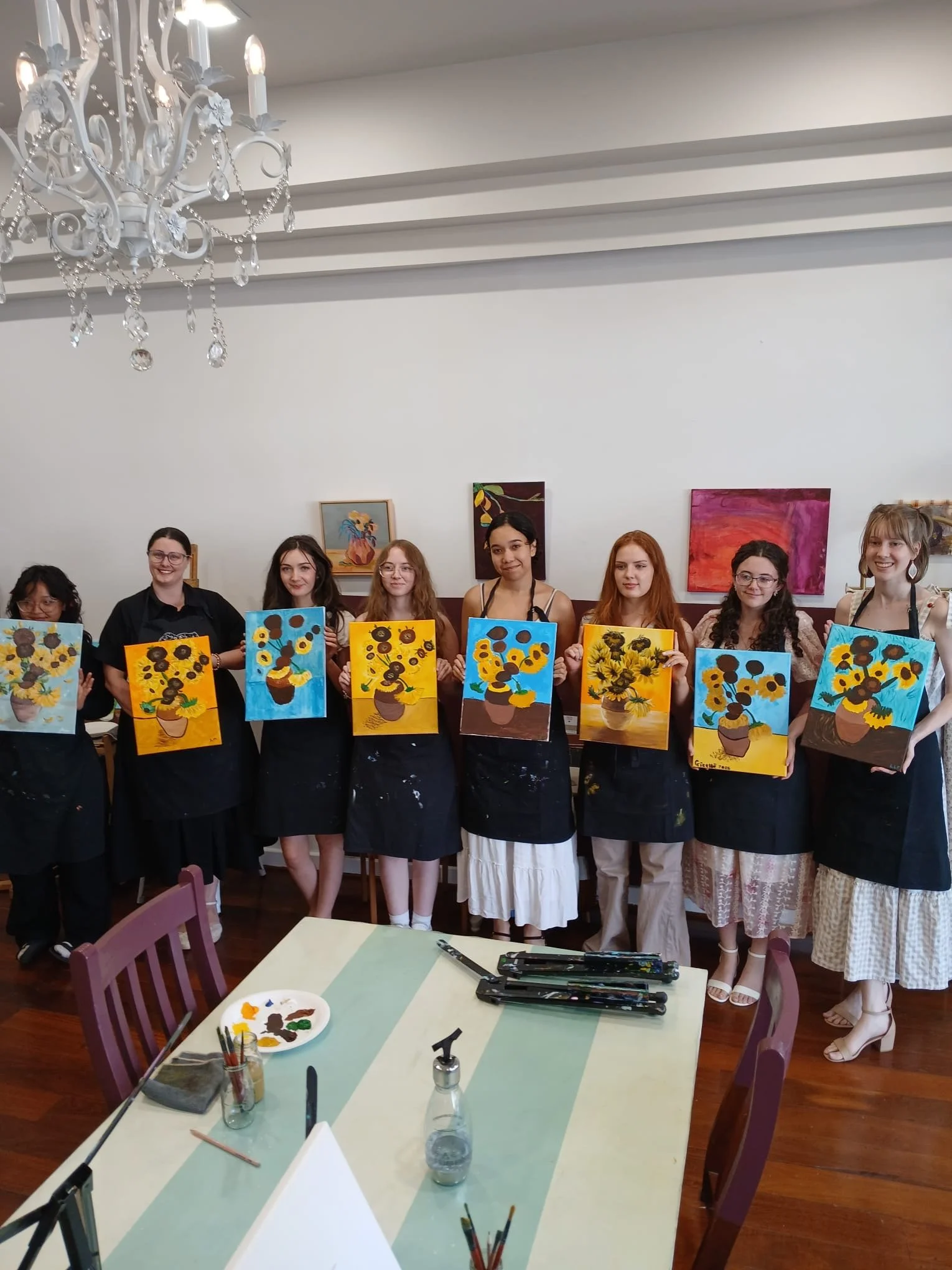 Group of women standing in a row, holding paintings of sunflower still life, inside a room with white walls and artwork on the wall, in front of a table with art supplies.