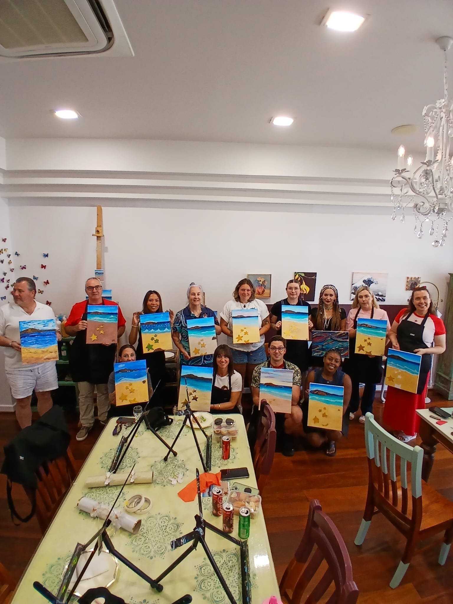 Group of people holding up paintings of a beach scene with sand, ocean, and sky, in a room with a chandelier and artwork on the wall.