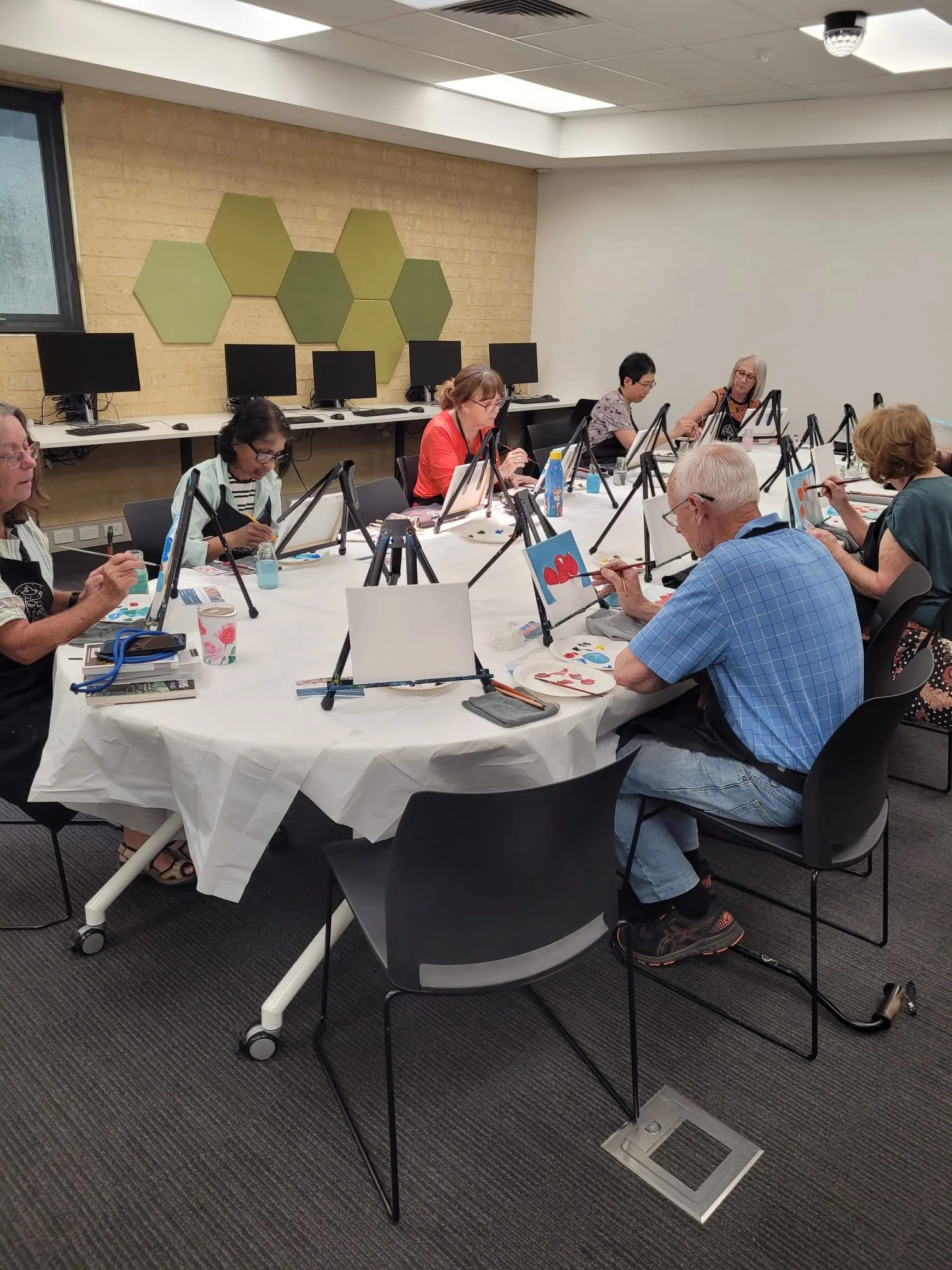 A group of people sitting around a large table, painting on canvases that are propped on easels during a painting class in a room with computers and hexagonal wall decorations.