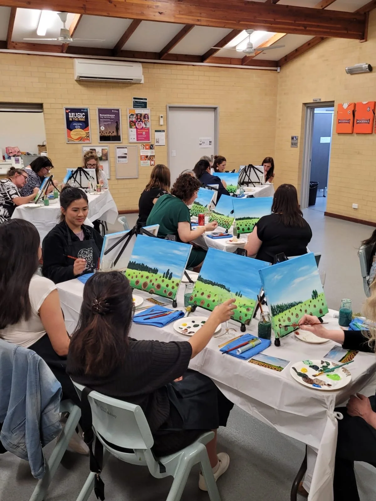 People participating in a painting class, working on landscape paintings of green fields and blue skies, seated at long tables with art supplies in a bright room with brick walls and wooden beams.