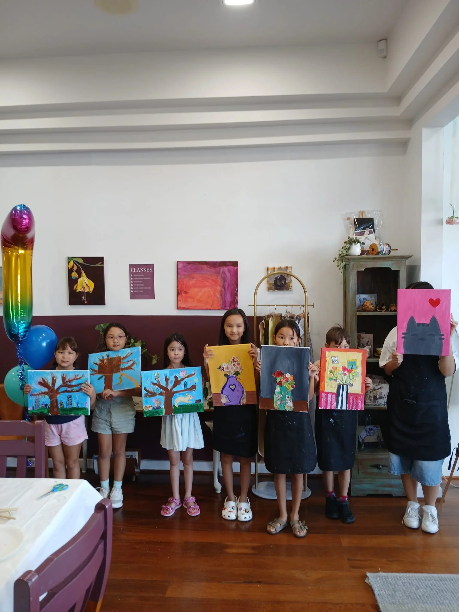 Seven children standing indoors, each holding up colorful paintings of various subjects, with some children dressed casually and some in aprons, celebrating an art event or class.