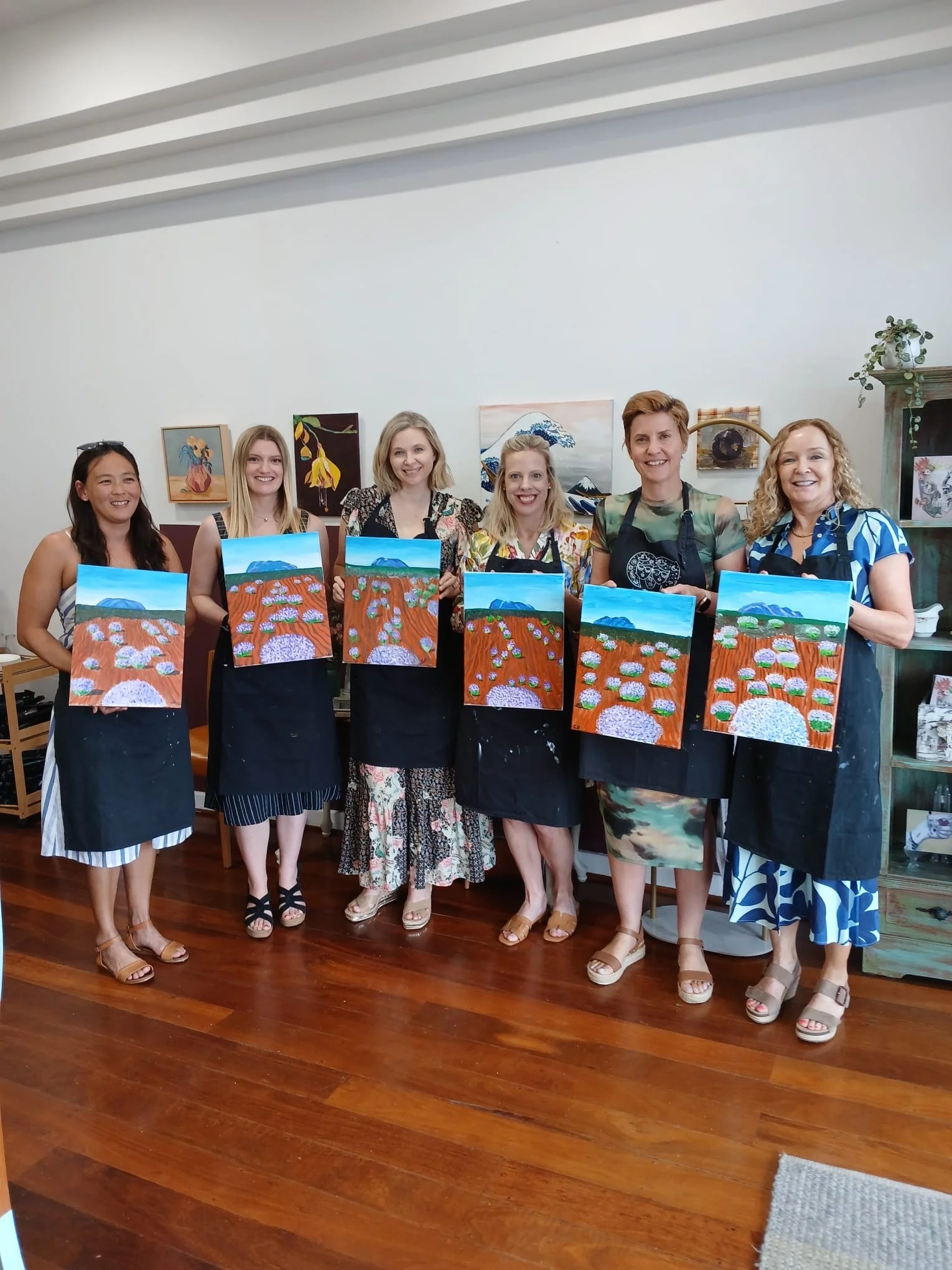 Group of seven women at an art class, holding their paintings of a landscape with orange flowers and blue mountains, standing in a gallery with wall art behind them.