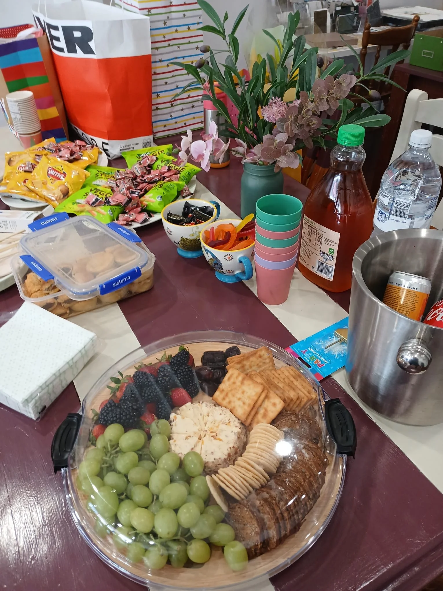 A table set with a large fruit and snack platter, colorful cups, snacks, drinks like juice and soda, paper napkins, and decorations including potted plants and wrapping paper in the background, suggesting a casual gathering or party.
