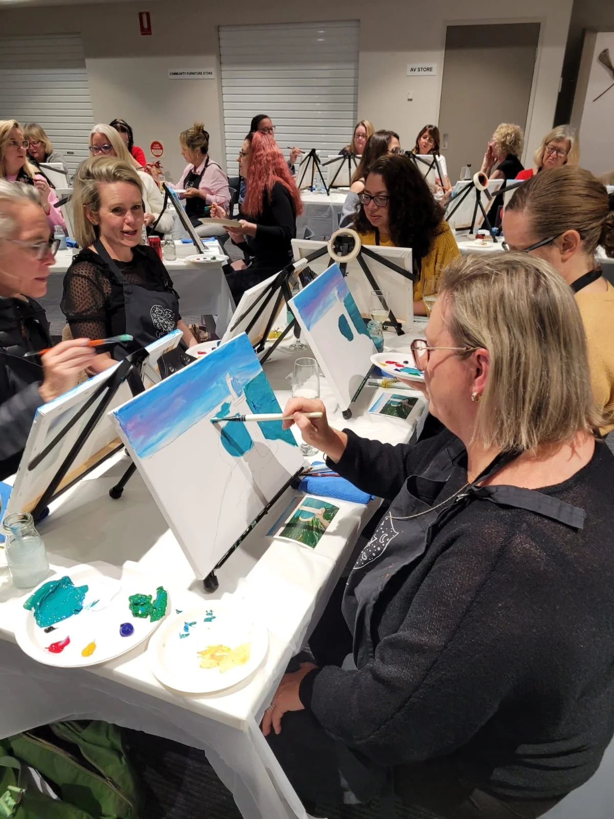 Group of women participating in a painting class, sitting at tables with canvases, paints, and brushes, in a well-lit indoor setting.